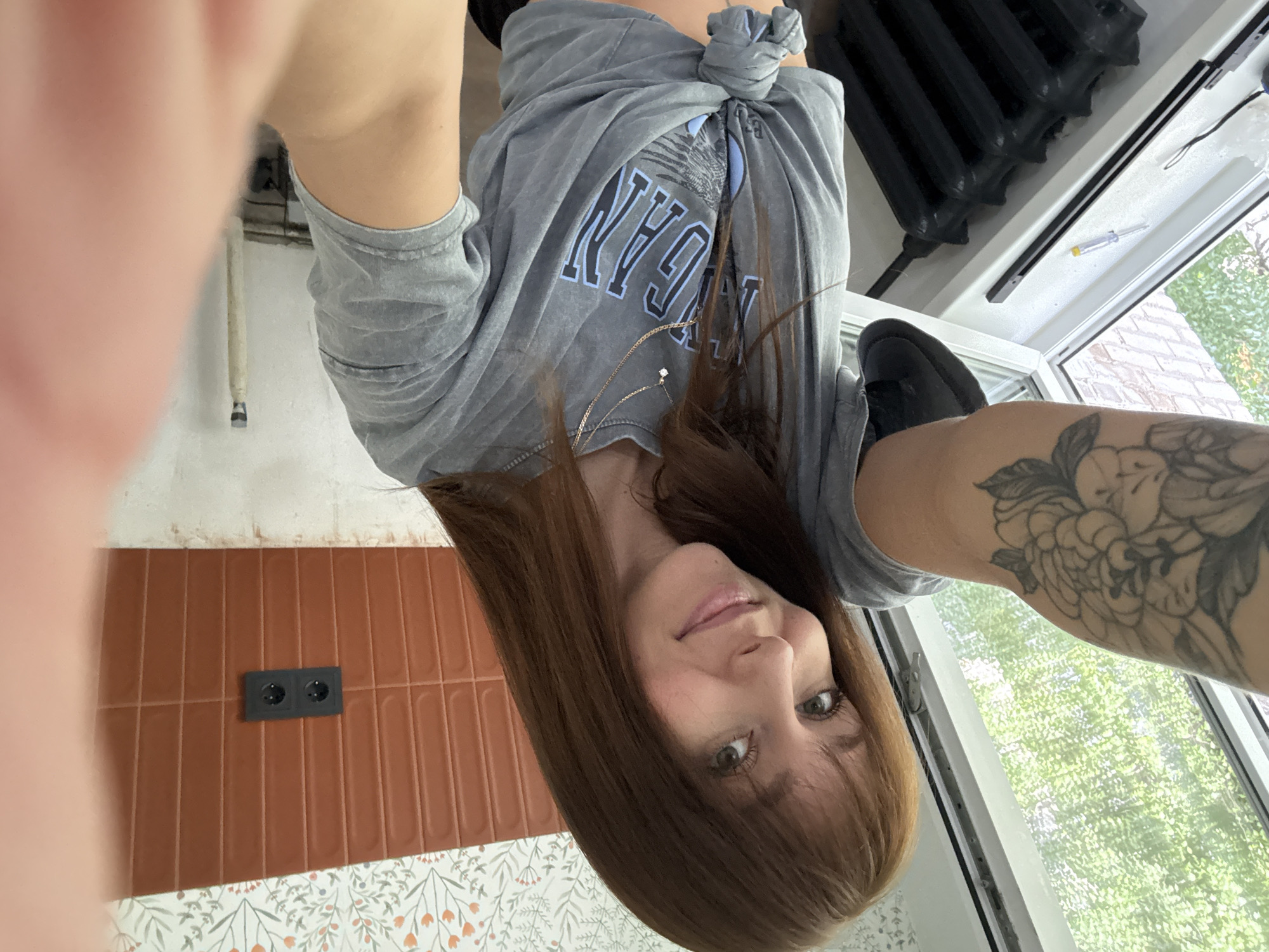 The media file is a selfie taken by a young woman indoors, with a partial view of a room. She has long, brown hair and appears to be looking at the camera with a slight smile. She is wearing a grey t-shirt, knotted at the front, with a blue graphic design and text that reads "YANKEE" above what looks like "NY". A delicate gold chain with a small pendant is visible around her neck.
To her left, there is a wall with two electrical outlets set into it, one above the other. The wall is partially covered with rectangular, reddish-brown tiles, and below that is wallpaper with a floral pattern in white and green. To her right, a large window shows a view of green trees outside, suggesting it is daytime and the weather is fair. A black radiator is visible along the windowsill. Part of her left arm, with a tattoo of flowers, and her right leg are also visible. The overall impression is of a casual, personal snapshot.