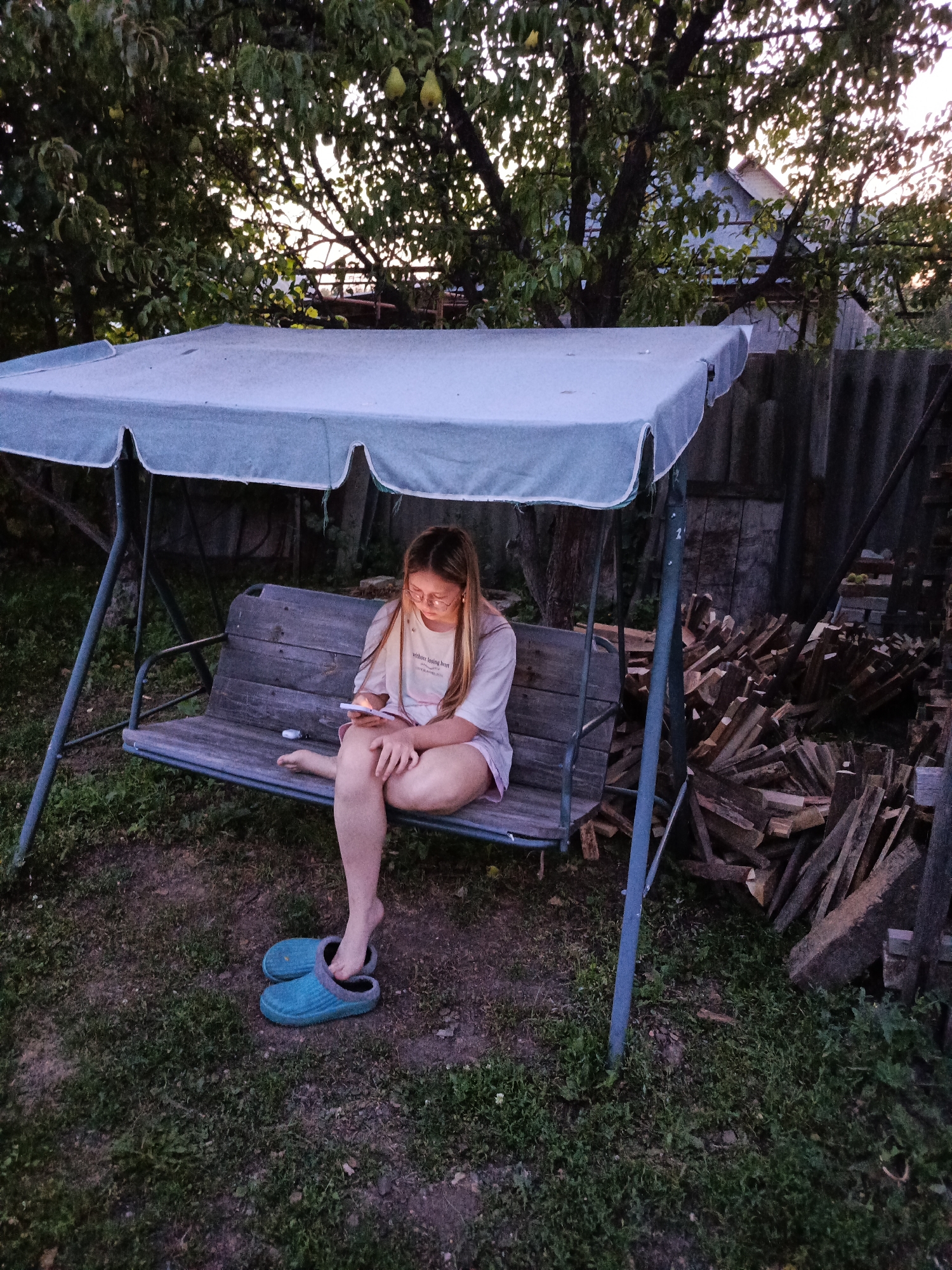 In a backyard setting, a young woman is seated on a wooden swing set bench, engrossed in her smartphone. She is wearing a light-colored t-shirt with text and pink shorts, and has blue slip-on shoes on her feet, one of which is partially off. The swing set is covered by a light blue canopy. Behind the swing set, a pile of cut firewood is stacked next to a corrugated fence. Trees with visible fruit are in the background, and a house is partially visible through the foliage. The scene appears to be taking place in the late afternoon or early evening, given the lighting conditions. The image is believed to have been taken in Saraktash, Russia, though this cannot be definitively confirmed by the visual information alone.
