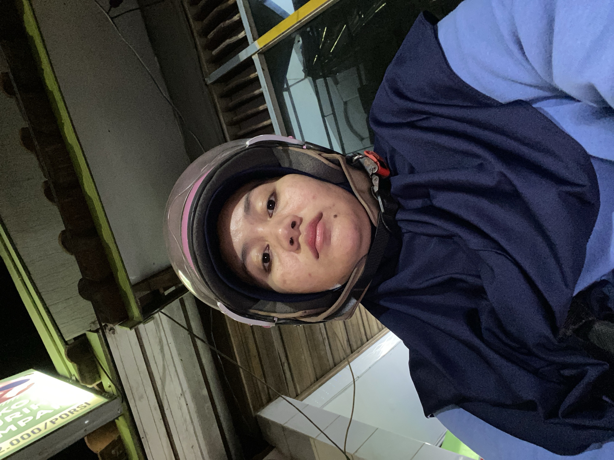 A portrait of a woman wearing a helmet and a dark blue hijab is shown from a low angle. She is looking directly at the camera, her expression neutral. The background consists of a building's exterior with wooden slats and a bright, illuminated sign with Indonesian text. The lighting suggests it is nighttime or dusk. The overall environment appears to be an urban setting, possibly a street or an outdoor commercial area in Banjarbaru, Indonesia.