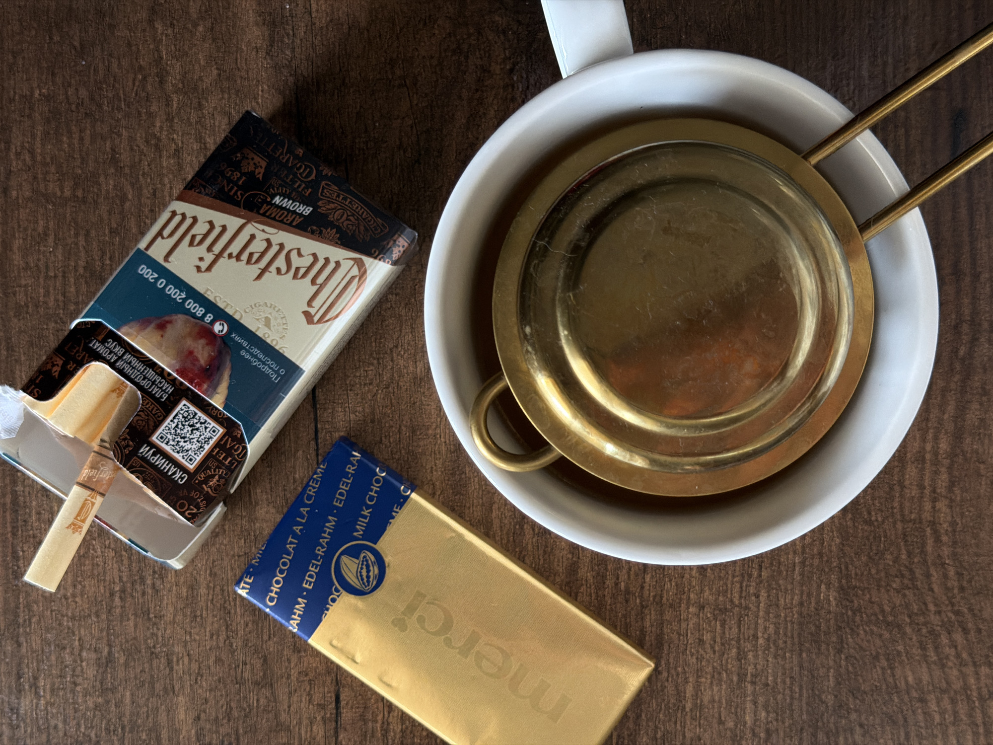 The image shows a still life arrangement on a dark wooden surface. A partially open pack of "Chesterfield" cigarettes sits to the left, with a cigarette partially extending from the pack. Next to it is a gold-wrapped "Merci" chocolate bar. To the right, a white mug contains a gold-colored tea strainer with handles. The setting could be in Rossiyskiy, Russia, based on the Cyrillic text on the cigarette pack.