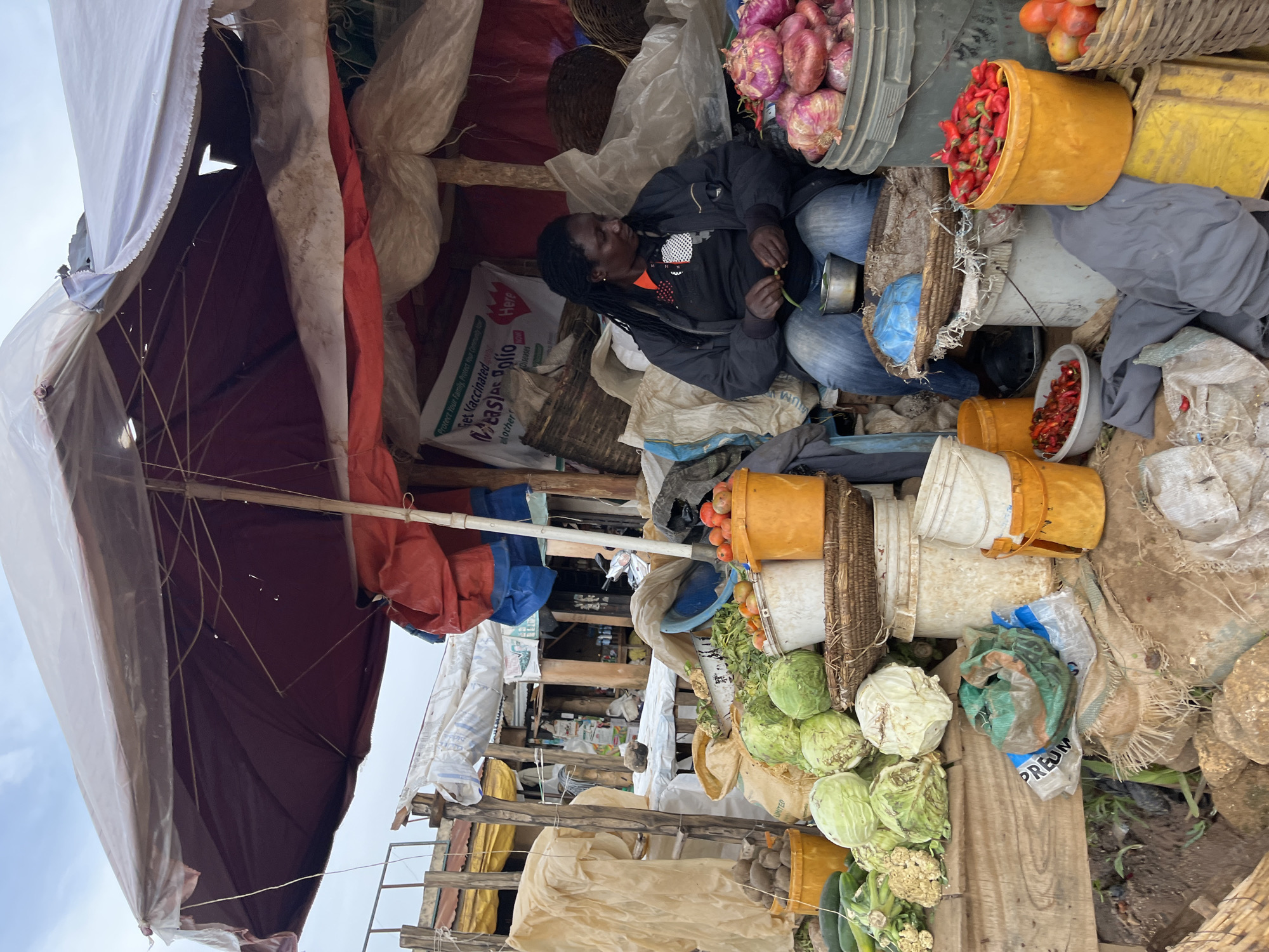 This media file depicts an outdoor market scene in Gura Top, Nigeria, likely during the daytime given the overhead natural light. The primary subject is a person, appearing to be a vendor, resting amidst a variety of produce and market goods. They are seated, wearing a dark jacket and blue jeans, with their eyes closed and a small green vegetable in their hands, suggesting they may be preparing produce or taking a break.
The setting is a bustling marketplace with an abundance of items for sale. Visible produce includes red onions, red chili peppers piled in buckets, tomatoes, cabbages, cauliflower, and cucumbers. Various containers like yellow and white buckets, baskets, and woven sacks are used to store and display the goods. The market structure is rudimentary, with visible wooden beams and coverings made of tarpaulin and fabric, some of which are torn or weathered. Bags of what appear to be grains or other goods are stacked around.
The overall impression is one of a typical day at a local market, with the vendor taking a moment of repose between sales. The sky is visible in the upper left, appearing overcast or hazy. Text visible on a white bag includes "Get Vaccinated," "Protect Your Family, Protect Your Community," and partially obscured words that may indicate brand names or product information, such as "PREUM" and "LIMITED." The scene conveys a sense of everyday commerce and community activity.