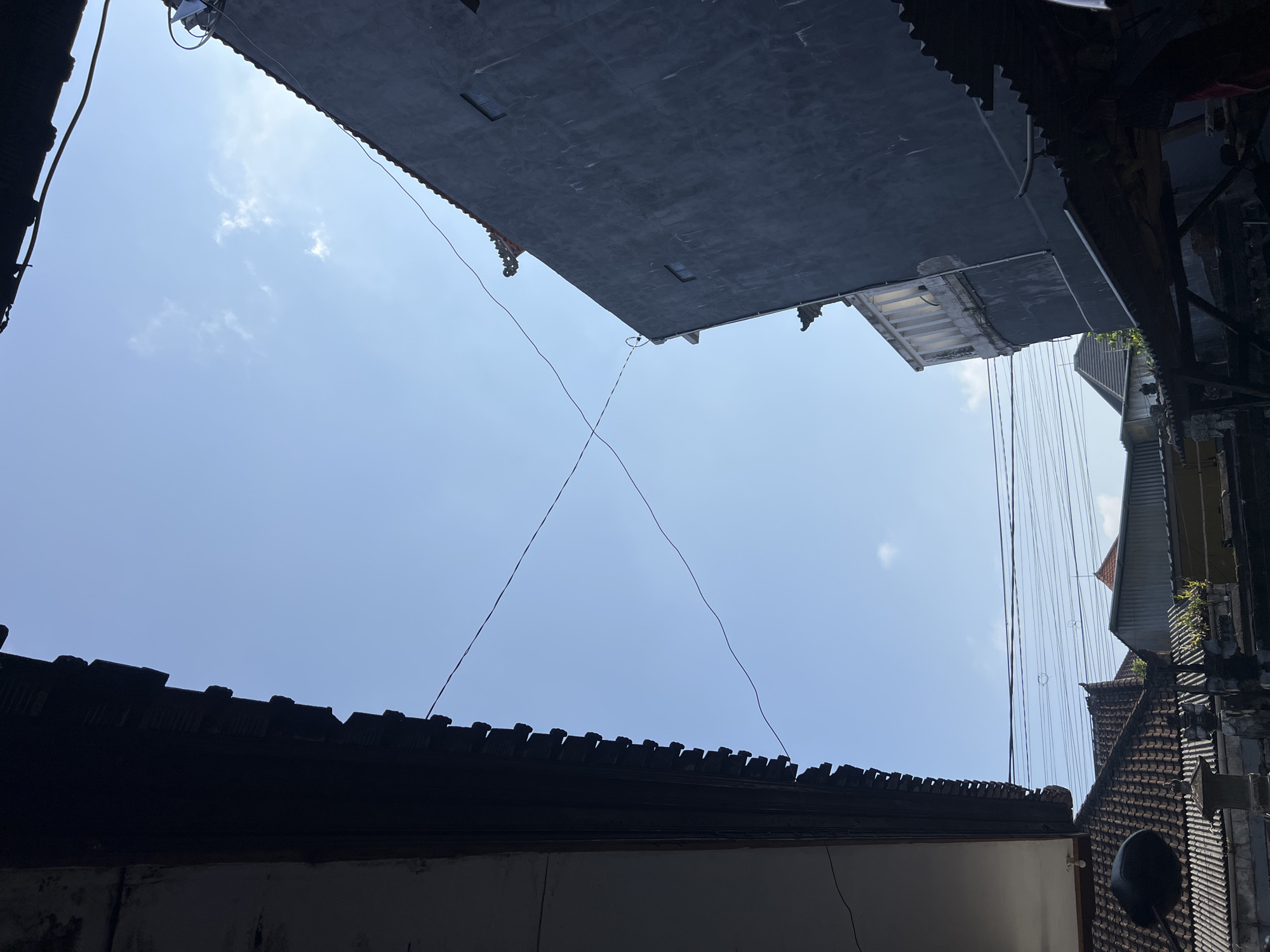 The image depicts an outdoor scene from a low-angle perspective, looking upwards towards a clear blue sky. The foreground is dominated by the eaves of two buildings, one on the left and another on the right, casting shadows. The building on the left has dark, layered eaves, while the building on the right is a tall, dark grey structure with visible windows and some decorative architectural elements at its peak. Thin wires crisscross the sky between these buildings. Below the eaves, the rooftops of several smaller buildings are visible, characterized by terracotta tiles and corrugated metal. The environment suggests an urban or semi-urban setting, likely in Denpasar, Indonesia, given the architectural style of the rooftops and the presence of overhead wires, which are common in many Indonesian cities. No people are visible in the image. The lighting indicates it is daytime, with the sun likely high in the sky, creating a bright and somewhat high-contrast scene.