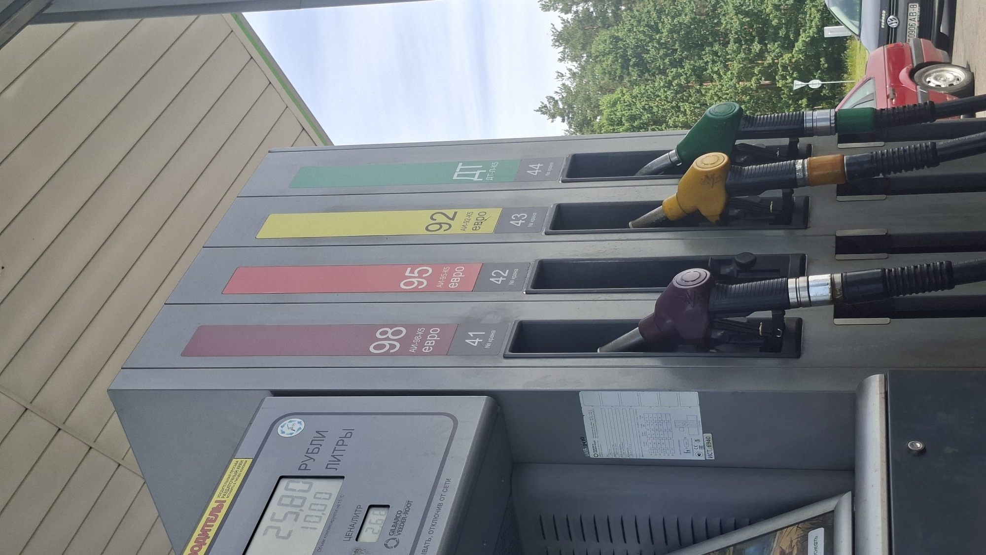 This media file shows a close-up of a gasoline pump at a service station. The pump has four different fuel nozzles, each labeled with the octane rating and type of fuel: 98, 95, 92, and DT-L-K5. The prices are displayed on the pump's digital screen, showing 25.80 rubles for volume and 2.68 for price per liter. The context indicates the location is in the Drachkovskiy rural council, Belarus.
The scene appears to be outdoors, with natural light suggesting daytime. The sky is visible in the background, partially obscured by trees. There are vehicles in the background, including a red car and another partially visible car. The overall setting is a typical gas station forecourt.
The focus is on the fuel pump and its display, with the nozzles and price information being the primary subjects. There are no people clearly visible in the frame, but the presence of the gas pump and vehicles implies a transaction or refueling activity is taking place or has recently occurred. The weather appears to be fair. The visible text includes fuel types, octane ratings, prices, and manufacturer information on the pump.