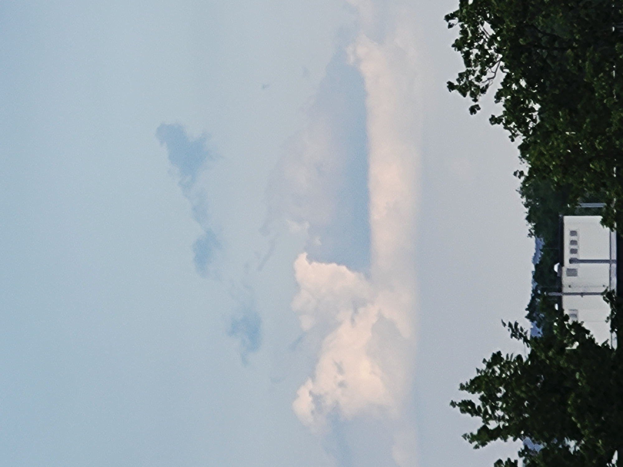 The image shows a view of a building and trees against a cloudy sky. The building is a light-colored, rectangular structure with multiple windows or openings. It is partially obscured by the dense green foliage of trees in the foreground. The trees are tall and leafy, with some branches extending into the sky. The sky is filled with a mixture of light and dark clouds, with some patches of blue sky visible. The overall impression is one of a somewhat overcast day with a mix of natural and man-made elements. The location is identified as Unknown, Unknown.