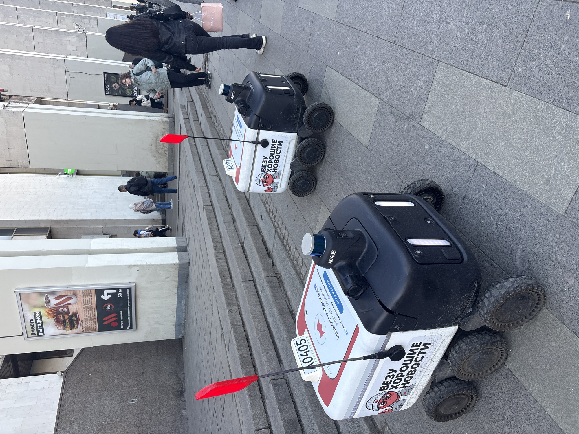 The image shows two Yandex delivery robots on a stone-paved area in Moscow, Russia. The robots are white and black, with six wheels each, and a red flag attached to their rear. The front robot is labeled "A0405" and the rear robot "A0277". The robots have signage in Russian reading "ВЕЗУ ХОРОШИЕ НОВОСТИ" which translates to "I bring good news". The signage also includes information about subscribing to Moscow news.
In the background, there are people walking, a building, and a sign advertising food from "ВКУСНО И ТОЧКА" which translates to "Tasty and that's it". The sign mentions the establishment is 50m from the "здания Московского дворца молодежи" (Moscow Palace of Youth). The sign in the background reads "MOS 20%". There is also a Payot bag.