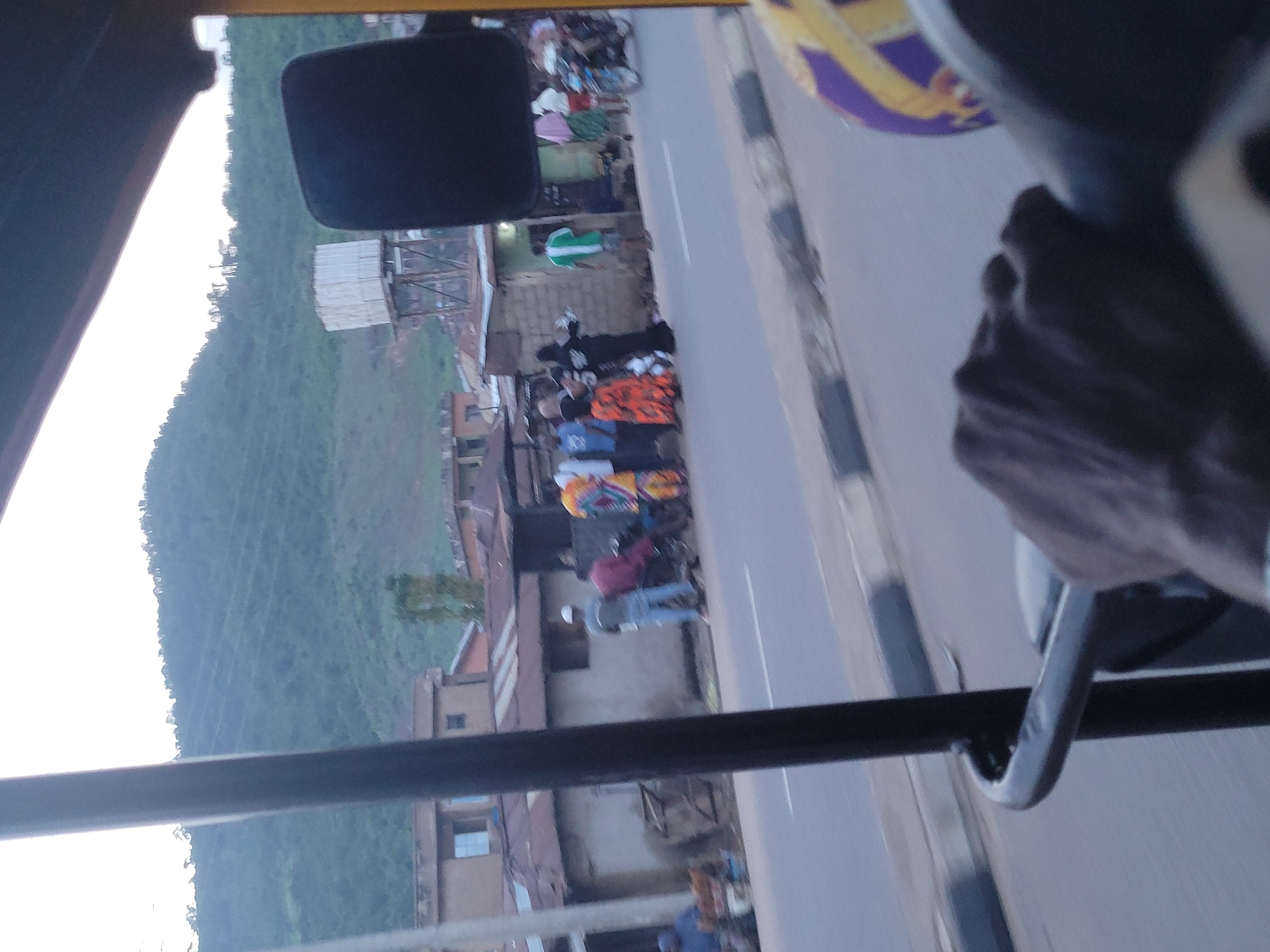 The image, taken in Lokoja, Nigeria, offers a glimpse of a street scene from what appears to be the interior of a vehicle. The perspective is tilted, suggesting the photo was taken from a moving vehicle. In the foreground, a hand is visible, gripping a metallic rod, possibly a grab handle within the vehicle.
The scene outside the vehicle is bustling. A group of people are gathered on the side of a paved road. Some are standing, while others are seated, possibly on motorcycles or scooters. The people are dressed in a variety of clothing, with some wearing bright colors. One individual is wearing a white cap and blue jeans, another a maroon top and blue jeans. There are also individuals in what appear to be traditional African attire with vibrant patterns.
Buildings line the road, some appearing to be simple structures with corrugated metal roofs, while others are more substantial, with windows and balconies. A green, grassy hillside rises behind the buildings, suggesting a somewhat hilly terrain. The lighting indicates it is daytime, likely late afternoon or early evening, as the light seems diffused and not harsh. The overall atmosphere appears to be that of a typical day in a busy Nigerian town. No visible text is present in the image.