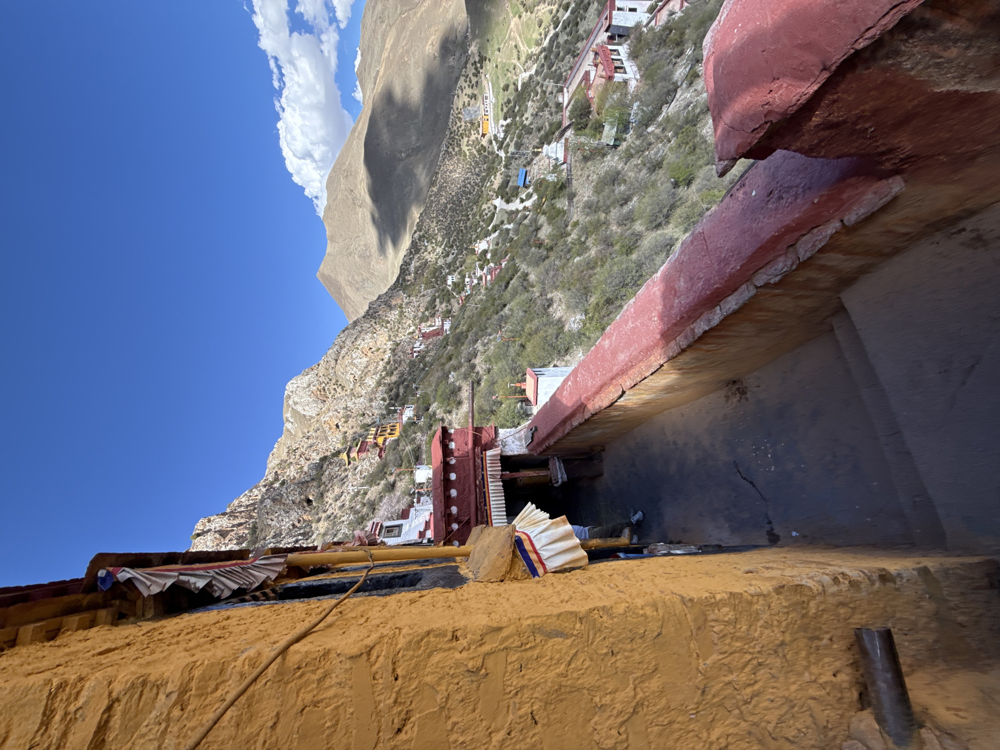 The image shows a view from what appears to be a high vantage point, likely within the city of Lhasa, China. The architecture is characteristic of Tibetan buildings, with textured yellow walls and red accents on the roofs. There are layers of buildings and structures visible, descending down a mountainside covered with vegetation. In the distance, a large mountain is partially obscured by clouds. The sky is a clear blue.
