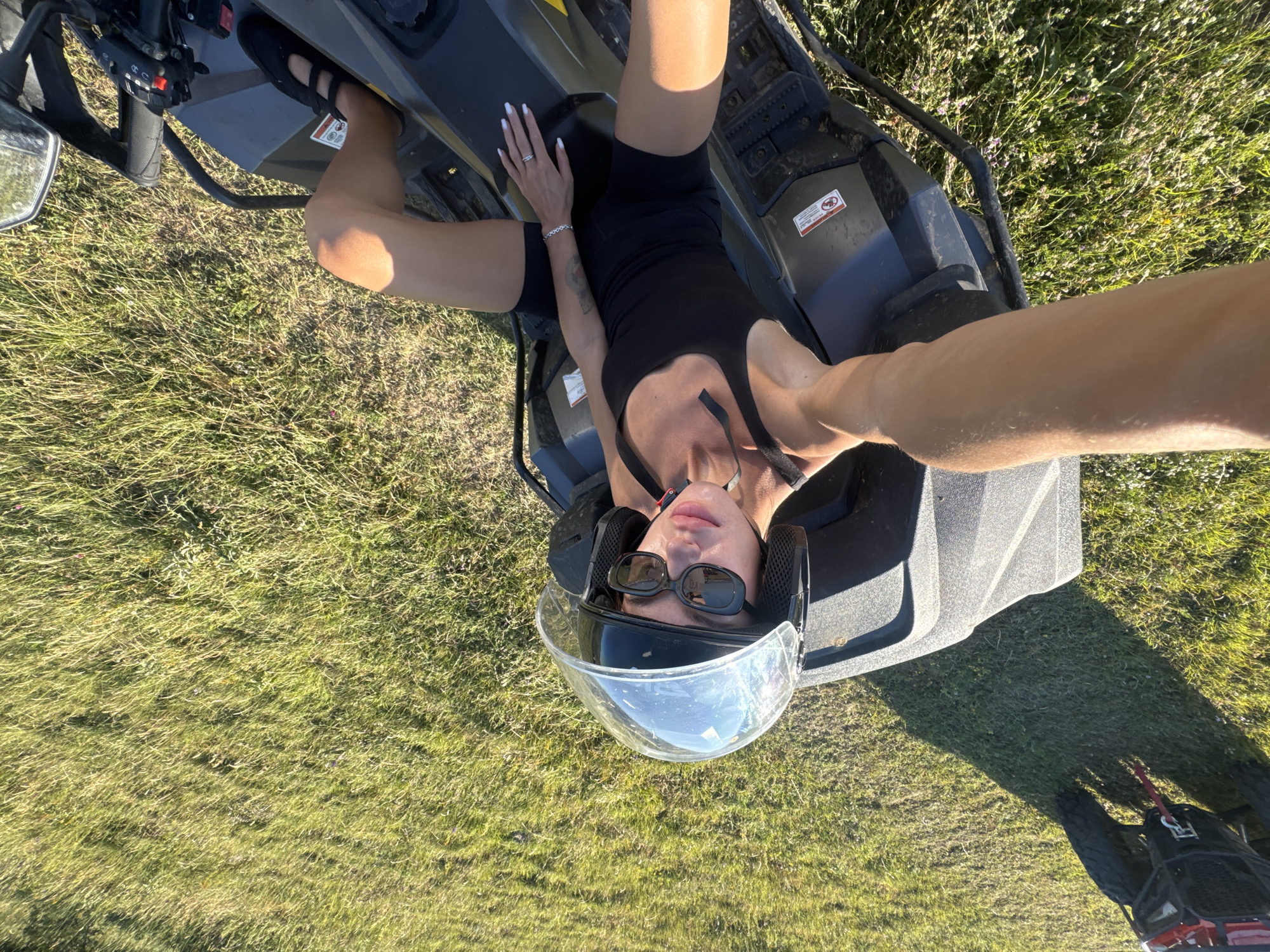 A woman wearing a black tank top, black shorts, sunglasses, and a helmet with the visor up is sitting on a dark grey ATV. She is lying back with her legs extended. Her right hand rests on the ATV, and her left hand is on her thigh. The ATV is parked on a grassy field. The image is taken from a high angle, with a person's arm extending into the frame from the bottom left. There is a shadow of the ATV cast on the grass. The background shows more green grass and some small purple flowers. The image is in color and appears to be taken during the day. There is no indication of the city of Tbilisi, Georgia, in the image.