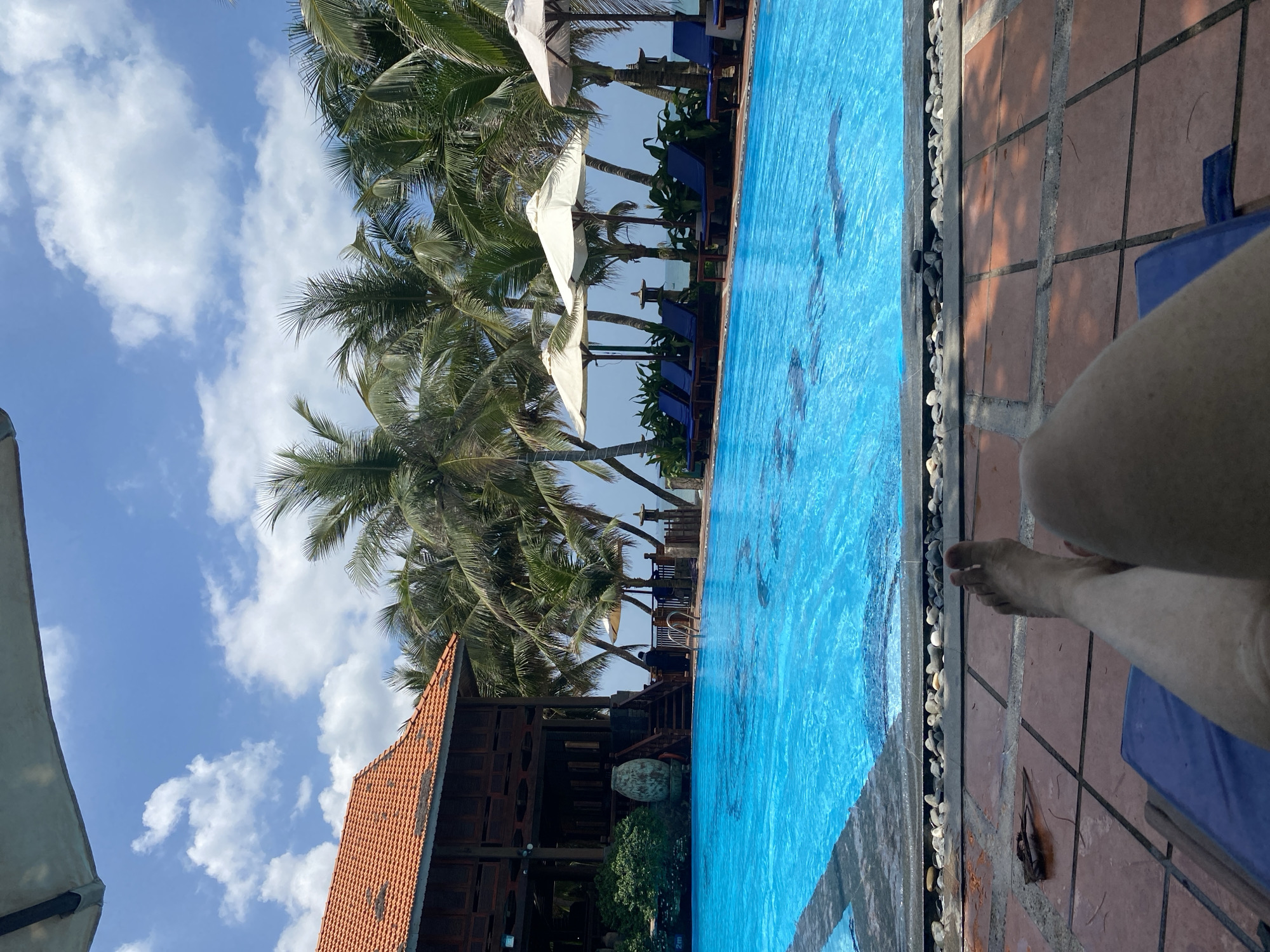 A person's legs and feet are visible in the foreground, resting on a blue lounge chair. The person appears to be in a relaxed position, facing away from the camera. The lounge chair is placed on a tiled patio area. Beyond the patio, a large, bright blue swimming pool dominates the mid-ground. The pool is surrounded by a stone border and palm trees. In the background, a large wooden structure with an orange tiled roof is visible, along with several white umbrellas providing shade for lounge chairs. The sky above is blue with scattered white clouds. The overall scene suggests a tropical resort setting in Phu Thuy Ward, Vietnam, likely during daytime.