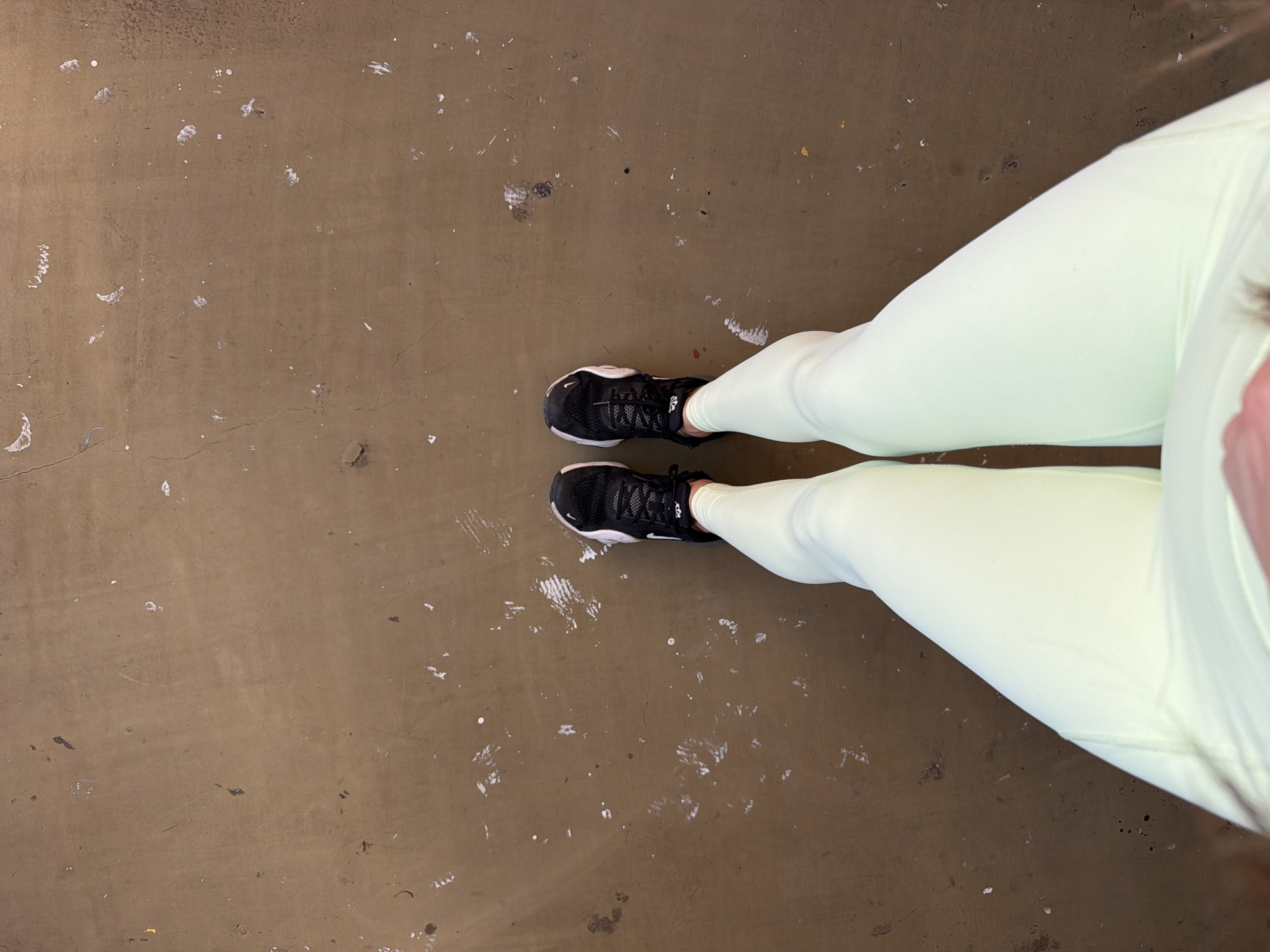 A top-down, eye-level shot shows a person standing on a dirty, beige-colored floor with many small white spots or splatters scattered across its surface. The person is wearing black athletic shoes with white soles and light green leggings. The shot includes the person's lower legs and feet, facing towards the center. The background is the floor, which seems to be in an indoor location. The setting appears to be utilitarian and possibly industrial. No context suggests the city of Sofia, Bulgaria.