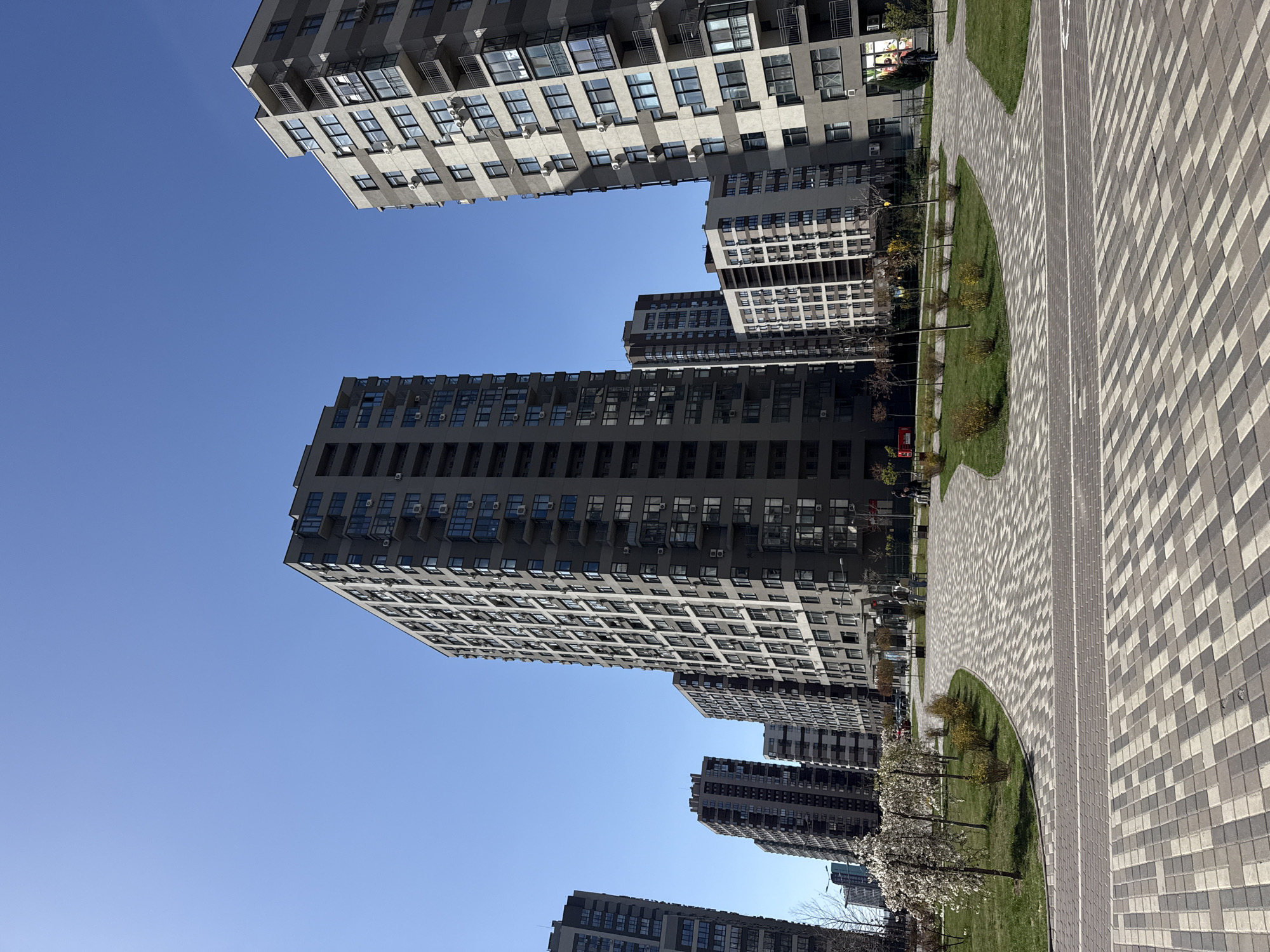 The image depicts a series of modern high-rise apartment buildings in Kyiv, Ukraine. The buildings feature a grid-like pattern of windows and balconies. The sky is clear and blue. A pedestrian walkway with gray and white patterned paving stones curves through a green area. Small trees and shrubs line the walkway. There are no visible people or interactions occurring in the scene.