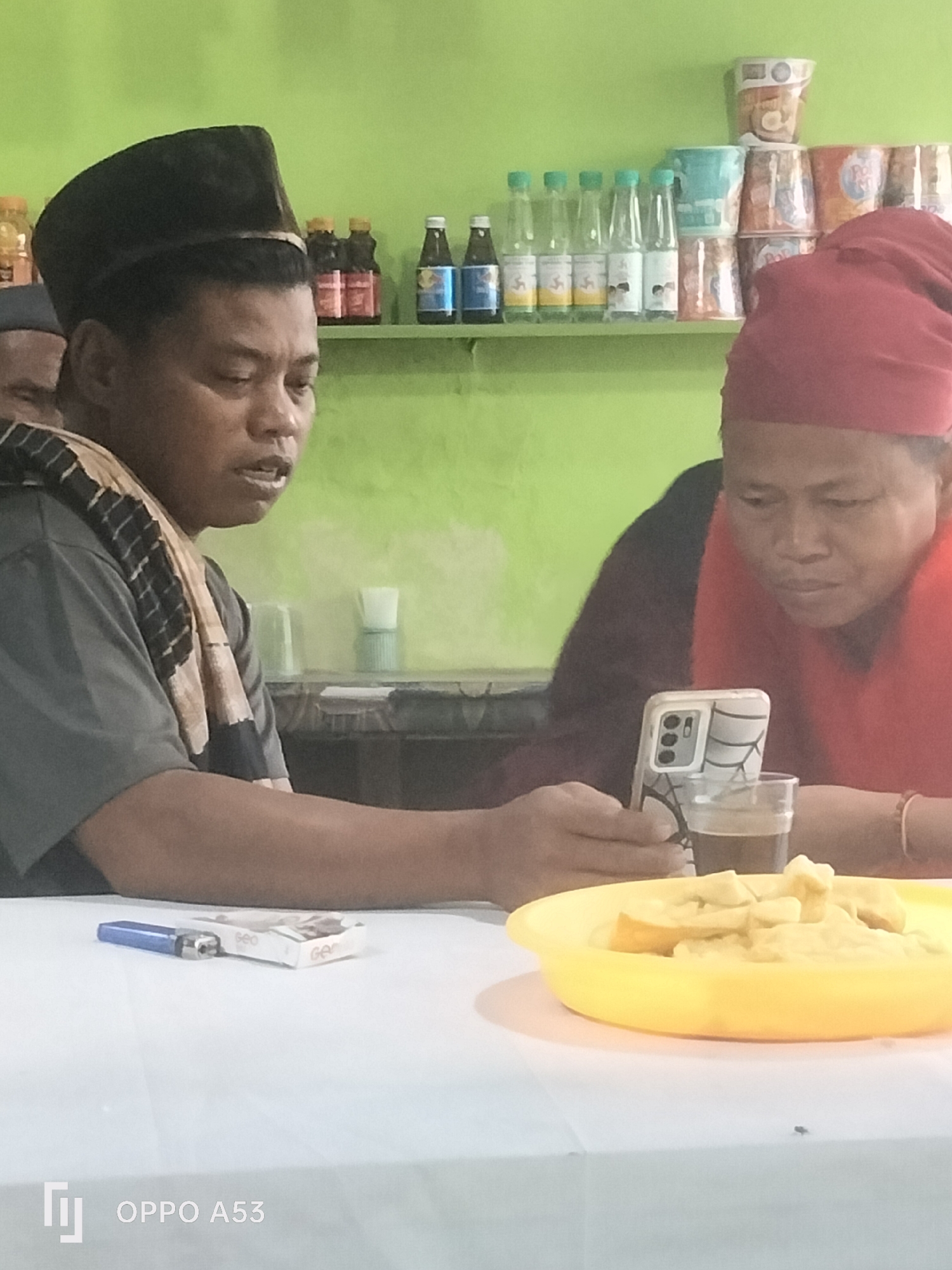 The image depicts two individuals seated at a table, likely in a food stall or local eatery in Badung, Indonesia. On the left, a person wearing a black peci hat and a patterned scarf is looking towards their right, with an open mouth as if speaking. In front of them is a cigarette pack and a blue lighter. To the right, another individual, wearing a red head covering, is focused on a smartphone held in their hands. The phone is propped up with a colorful case. Between the two individuals, on the table, is a small glass containing a dark liquid, possibly coffee, and a yellow plastic bowl filled with food items, which appear to be fried or battered. In the background, a shelf displays various bottles and packaged goods, consistent with a retail or food service setting. The wall behind the shelf is painted a vibrant green. The foreground is dominated by the white tablecloth covering the table. The overall scene suggests a casual interaction during a meal or break.