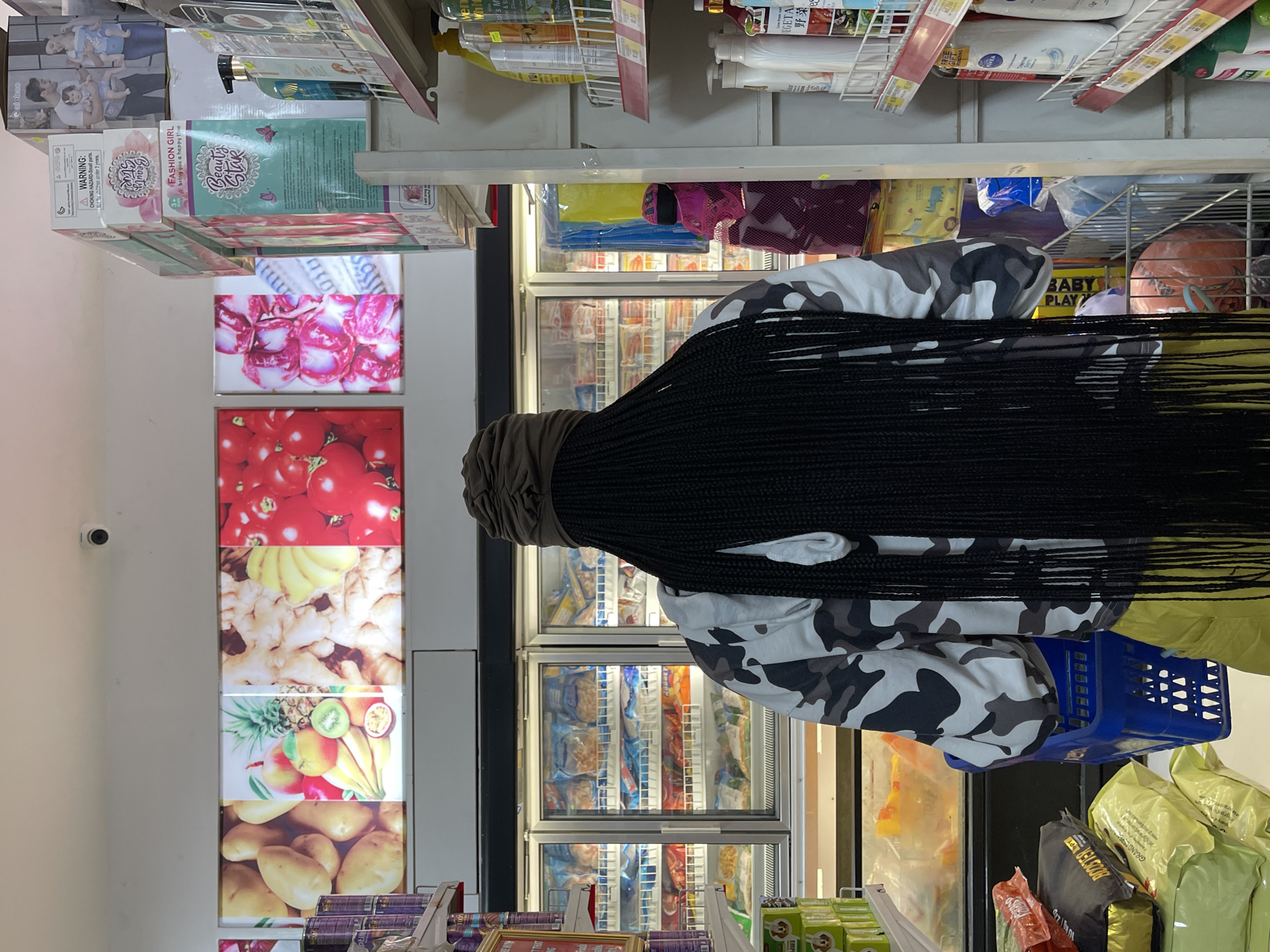 A person with long, black braided hair, wearing a camouflage jacket and a brown headwrap, stands in a brightly lit convenience store in Guzape, Nigeria. The person is facing away from the camera, holding a blue shopping basket. In the background, there are rows of refrigerated display cases filled with frozen food items. Above the display cases, a large illuminated advertisement features vibrant images of fruits and vegetables. To the right, shelves are stocked with various packaged goods, including bottles of cleaning supplies and personal care items. Stacks of boxed products are visible on higher shelves. The overall environment suggests a retail setting where groceries and household items are sold.