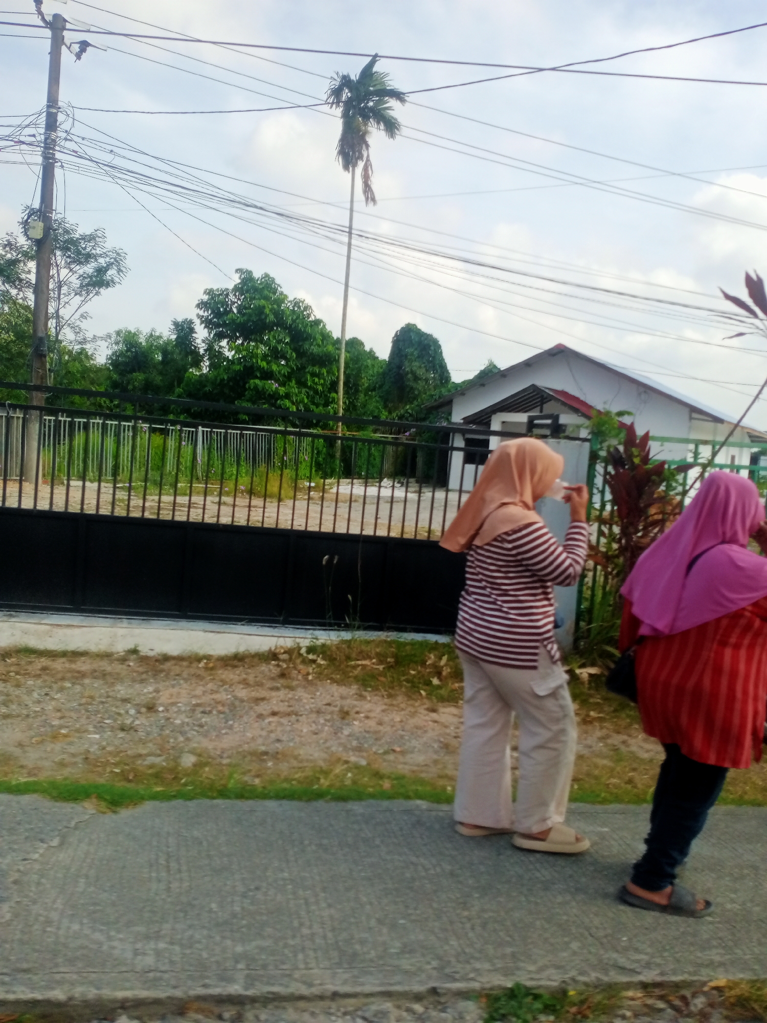 Two individuals wearing hijabs are walking on a paved sidewalk. The person on the left is wearing a striped long-sleeved shirt and light-colored pants, and is holding a transparent cup. The person on the right is wearing a red and black striped long-sleeved top and dark pants. A black metal fence with vertical bars runs along the background, with a white house visible behind it. Various trees and foliage are present, including a tall palm tree in the distance. Overhead, power lines crisscross the sky. The scene appears to be in a residential area, possibly in Balikpapan, Indonesia, given the presence of tropical vegetation and typical residential architecture. The lighting suggests it is daytime.
