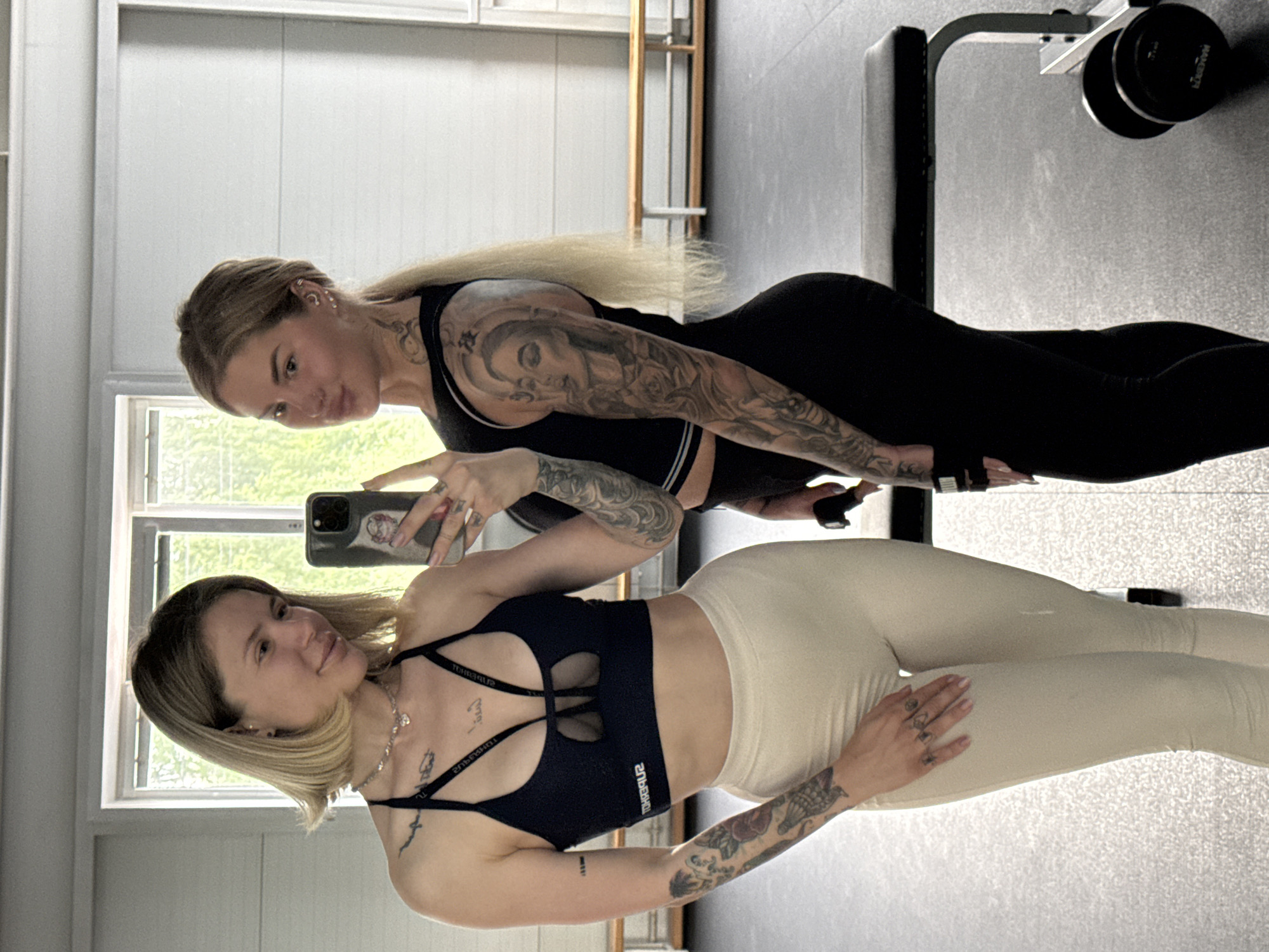 Two women are captured in a selfie taken indoors, likely a gym or fitness studio in Mayma, Russia. The image is a mirror selfie, showing the woman in the foreground holding a smartphone to capture the reflection of herself and her friend.
The woman closer to the camera has shoulder-length blonde hair, styled in a bob. She wears a dark blue or black sports bra with multiple straps and cutouts across the chest, featuring the reversed text "POWERLITE". Her lower body is clad in light-colored (possibly beige or off-white) leggings. She displays numerous tattoos on both arms and her chest. Her expression is a slight, natural smile.
Behind her stands another woman with long blonde hair. She is wearing a black sports top and black leggings, and has extensive tattoos on her right arm, forming a full sleeve, and visible on her neck/shoulder. Her expression is also a subtle smile.
The setting is an indoor space with light-colored paneled walls and large windows that reveal green foliage outside, suggesting daytime. In the background, gym equipment is visible, including a black workout bench with a metallic frame and a black dumbbell with partially visible text "MANG" (likely part of a brand name). The floor is a dark grey, possibly rubber matting common in gyms. Both women are dressed in athletic attire, indicating they are either working out, have finished, or are about to begin a session.