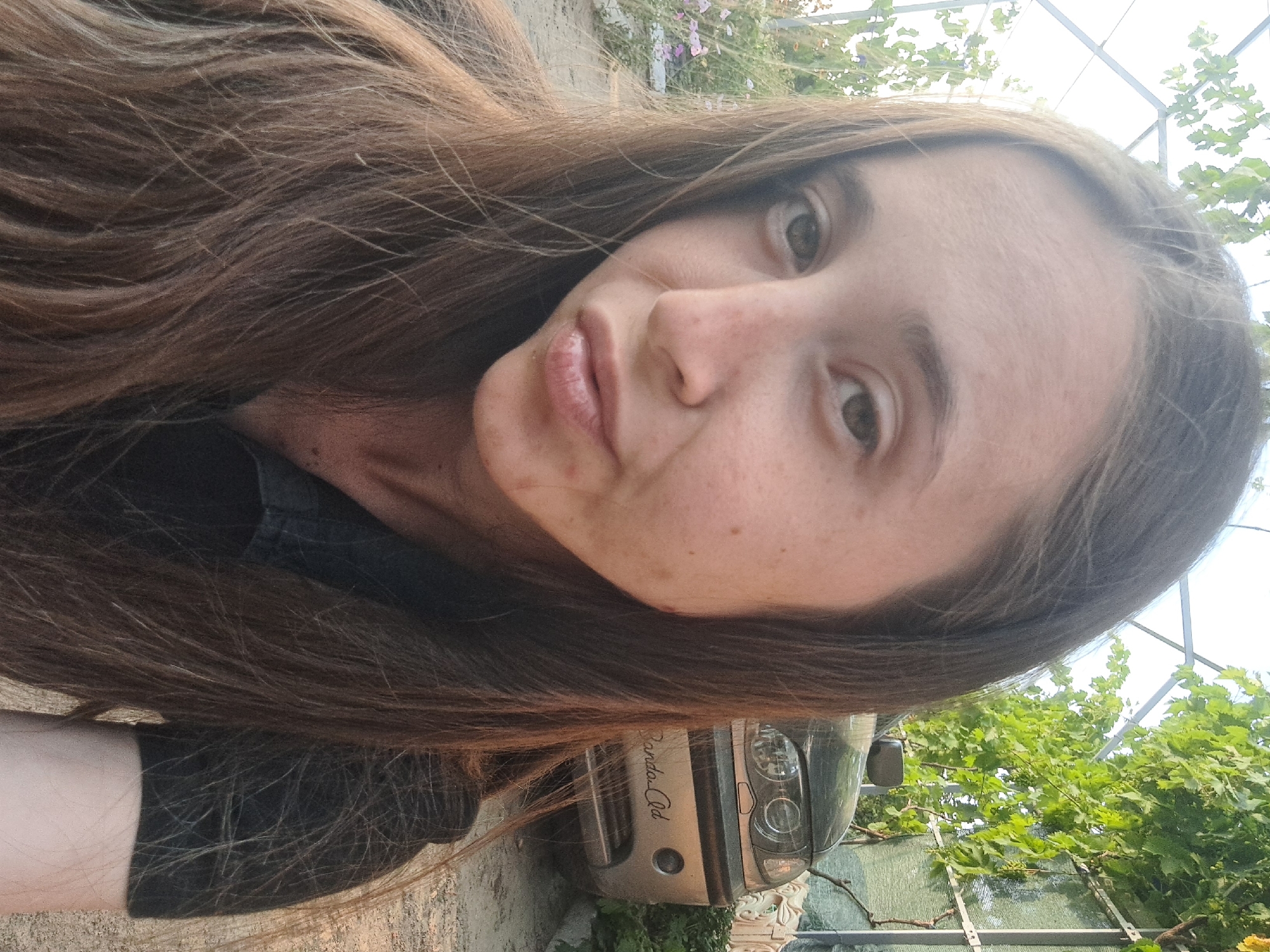 A young woman with long, brown hair and fair skin is captured in a close-up, selfie-style photograph. She is wearing a black shirt and her lips are slightly pursed. Her eyes are open and looking towards the camera. In the background, a silver car is visible with some text on its rear bumper. Green foliage and a white structure are also present, suggesting an outdoor setting. The ground appears to be dirt or gravel. The overall lighting indicates it is daytime. The location is stated to be in Vypasne, Ukraine.