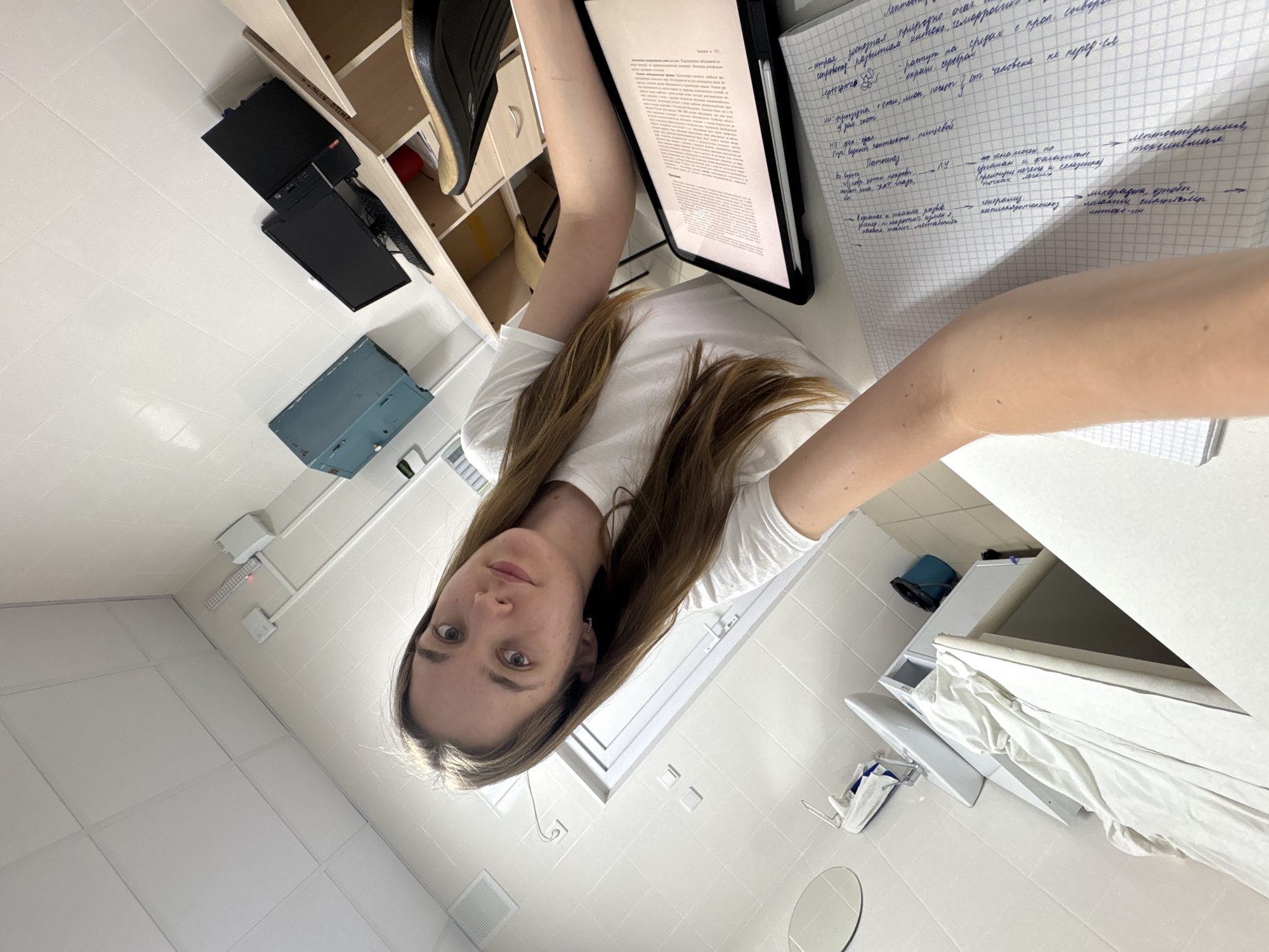 A young woman with long brown hair and a white t-shirt takes a selfie in what appears to be a study area or laboratory in Perm, Russia. She holds the camera, looking directly at it with a neutral expression. In front of her, on a white desk, an open tablet displays a document with text, and a grid-lined notebook contains extensive handwritten notes in Cyrillic script.
The visible text in the notebook, written in Russian, discusses medical or biological topics, with keywords such as "Патогенез" (Pathogenesis), "Лептоспиремия" (Leptospiremia), "токсинемия" (toxemia), "лихорадка" (fever), and "интокс-ли" (intoxication). Phrases like "от человека не передается" (not transmitted from human to human) are also discernible, suggesting a focus on infectious diseases or pathology. The tablet also shows text in Cyrillic, likely an academic or scientific article.
The room features white tiled walls and ceiling, indicative of a clean, possibly sterile, environment typical of medical or research facilities. To the left of the woman, a wooden shelving unit holds a computer tower, a monitor, a keyboard, and other items. Below the shelves, a blue-grey metal cabinet is visible. To her right, a white counter or table is seen, along with a white laundry hamper or cart containing white linens, and a blue bucket on a shelf. Electrical conduits and a ventilation grate are visible on the walls and ceiling, further suggesting an institutional setting.