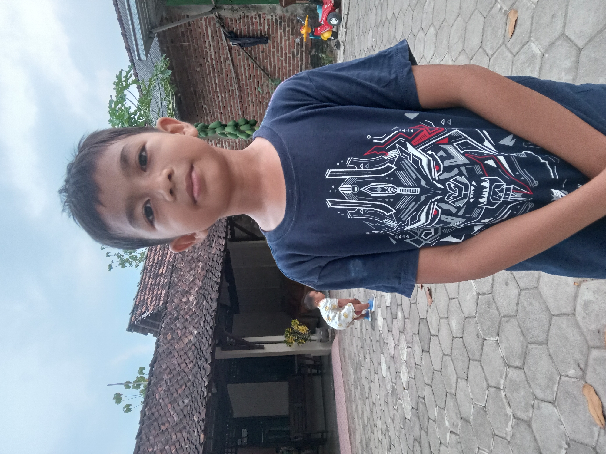 A young boy, appearing to be of Southeast Asian descent, stands in the foreground, facing the camera. He has dark hair and is wearing a dark blue t-shirt with a graphic design featuring a stylized, robotic-like face with red eyes. His hands are clasped in front of him. In the background, a younger child is bent over on a paved courtyard made of hexagonal stones. Behind the children, there is a traditional Indonesian house with a tiled roof and open verandas. A brick wall is visible on the right side of the image, with a cluster of green fruit hanging from a tree. A red and yellow toy ride-on vehicle is also present near the brick wall. The overall environment suggests a residential area in a rural or semi-urban setting, possibly in the village of Kedunggudel, Indonesia, given the architectural style and vegetation. The sky is overcast.