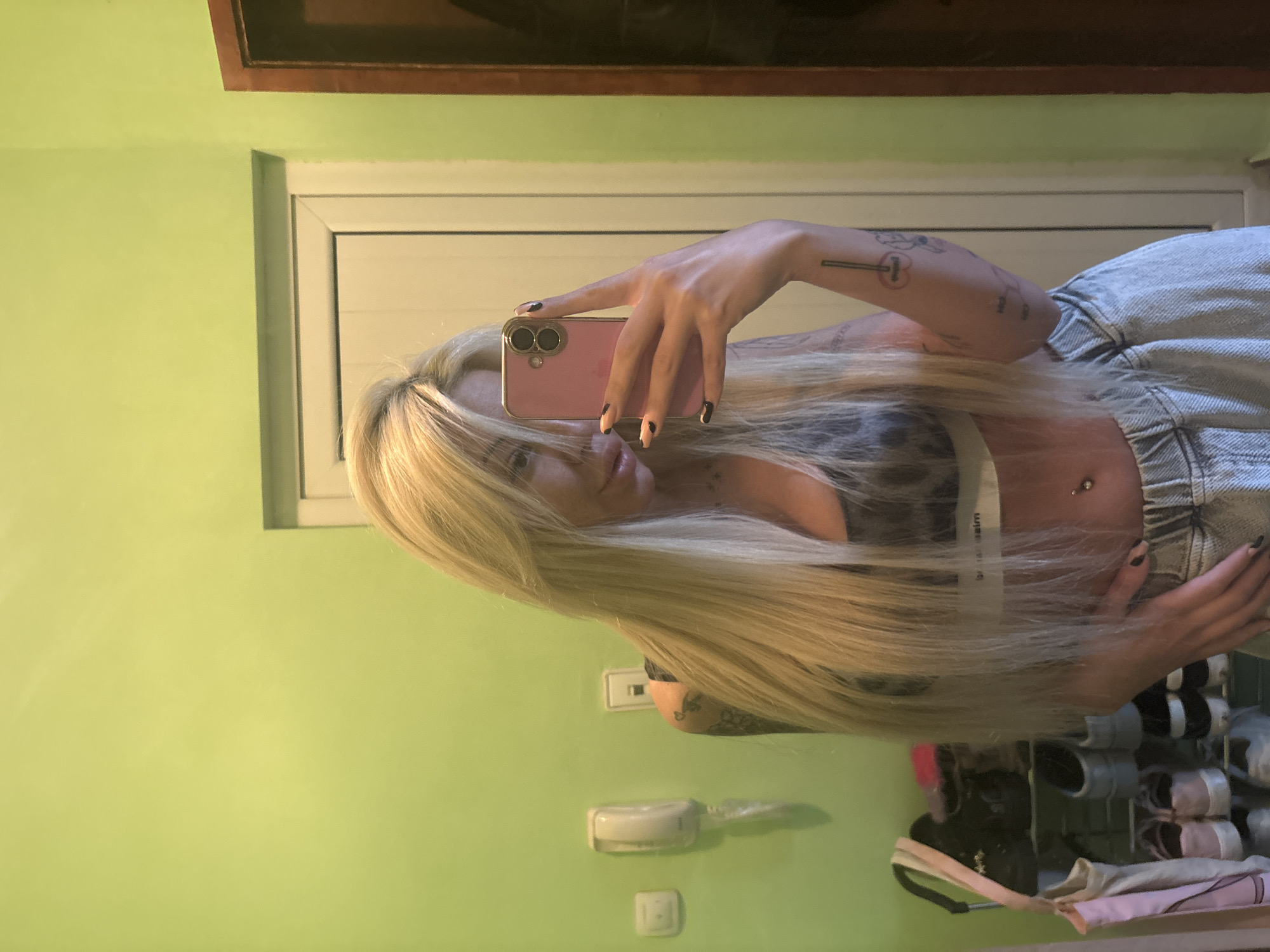 A woman is taking a selfie in front of a light green wall. She is holding a pink smartphone. The woman has long blonde hair that covers part of her face. She has a belly button piercing and is wearing a bra and jeans. The location is an interior space with a picture frame hanging on the wall above her and some accessories at the bottom corner. The city and location are unknown.