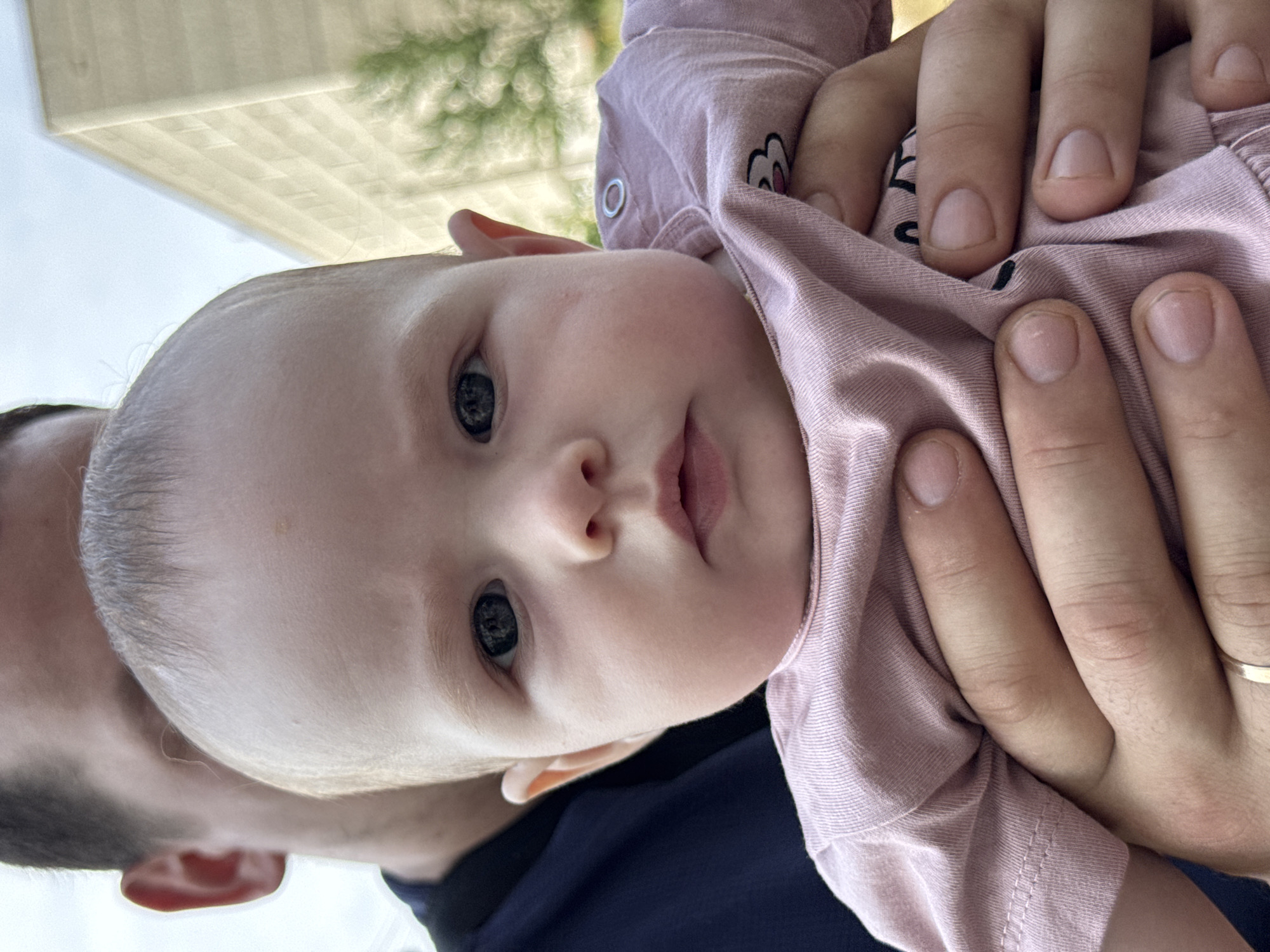 A close-up, medium shot features a baby being held by an adult in Noviy, Russia. The baby, positioned in the foreground and facing forward, has light skin, dark eyes, and light brown hair. The baby is wearing a pale pink t-shirt. The adult's hands are visible holding the baby, with a ring on one finger. The adult's dark-colored shirt is visible behind the baby. In the blurred background, a light-colored building and some greenery are present. The lighting suggests it is daytime.