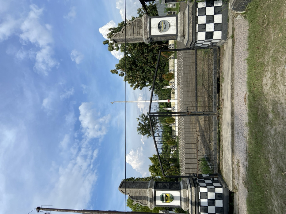 The image shows a closed black metal gate with checkered black and white concrete pillars on either side. The gate is located in Sukatani, Indonesia, at the coordinates -6.623, 107.404. Behind the gate is a paved area surrounded by a low fence with a large white pole in the center. Behind the pole are several green trees and a cloudy blue sky. The gate is likely part of a compound or a cemetery. There are no people present. The scene is quiet and peaceful.
