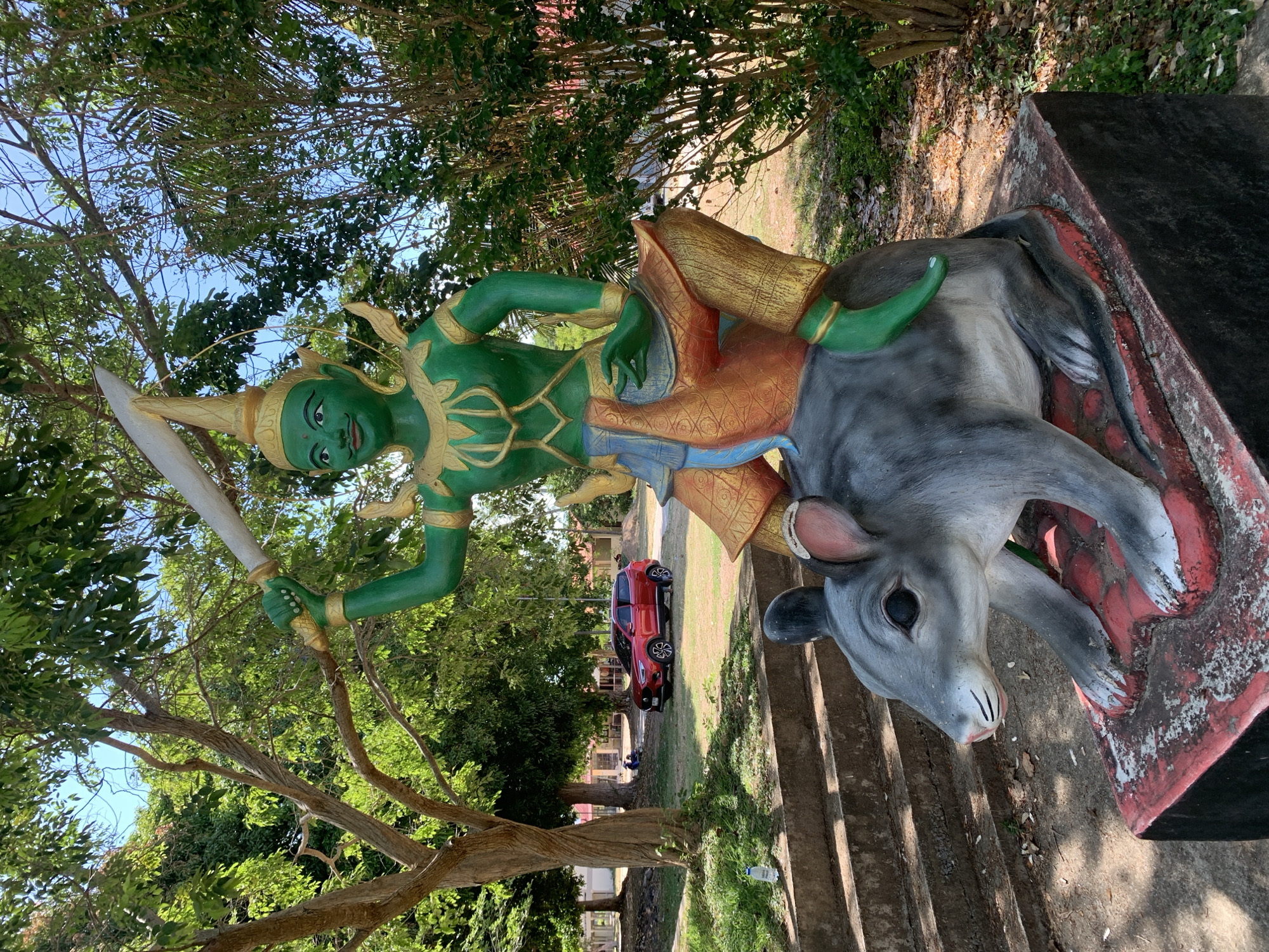 A statue of a green figure riding a gray ox stands in Baan Bo Phut, Thailand. The figure, clad in gold and orange, holds a sword aloft. A red car is visible in the background. The statue is situated beside concrete steps, with trees overhead.