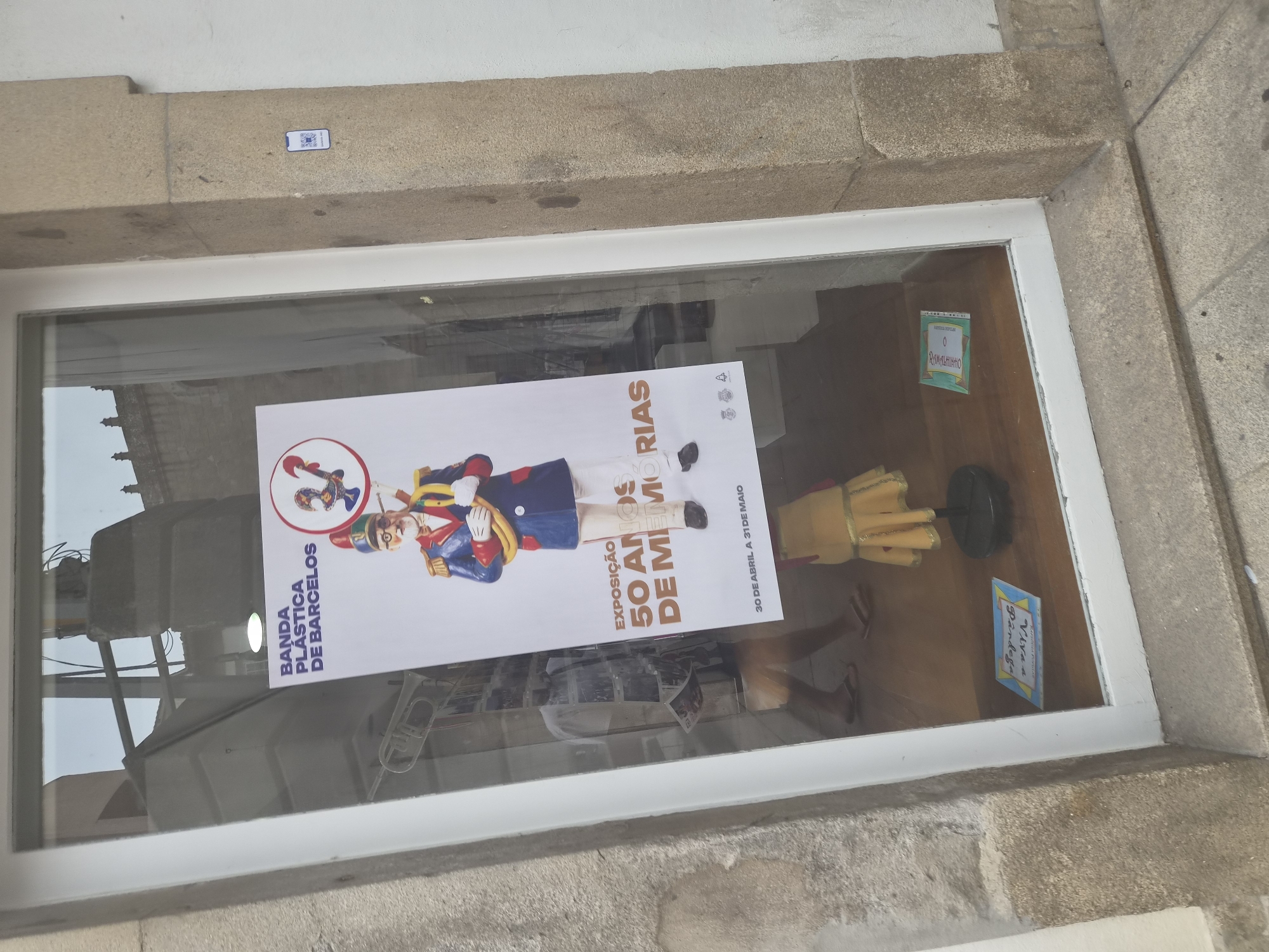 A poster for an exhibition is displayed in the window of a shop. The poster features a cartoon image of a man in a military uniform with a red hat and a trumpet. Text on the poster reads "BANDA PLÁSTICA DE BARCELOS" and "EXPOSIÇÃO 50 ANOS DE MEMÓRIAS". Below this, dates are listed as "30 DE ABRIL A 31 DE MAIO". The shop window also shows reflections of the street and people walking by. A QR code sticker is affixed to the stone pillar to the right of the window. The shop appears to be located in Barcelos, Portugal, as indicated by the poster's text.