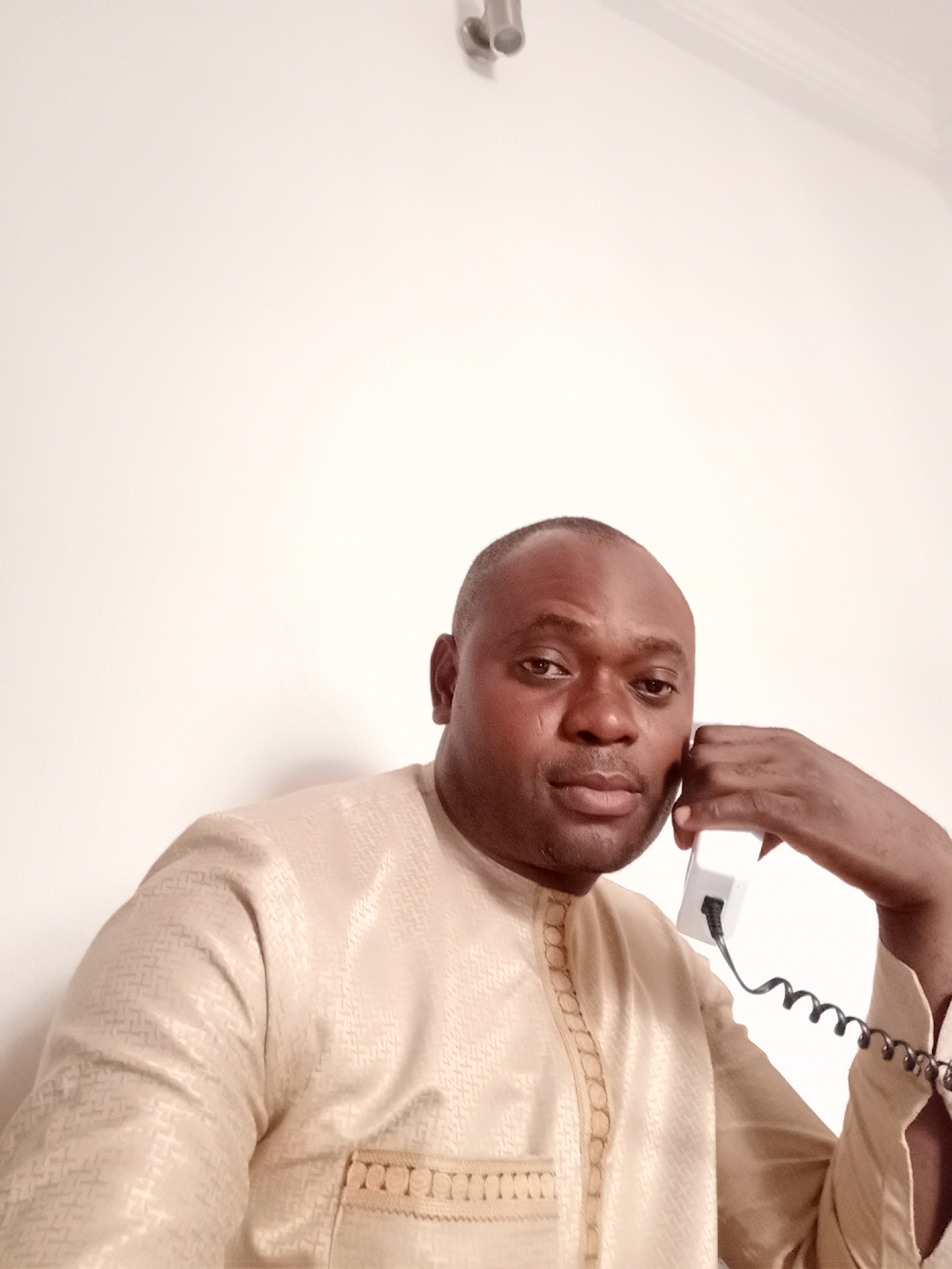 A medium shot of a man with dark skin and a shaved head is shown indoors. He is wearing a light tan, patterned shirt and holding a white telephone receiver to his left ear. The phone has a coiled black cord. The man is looking directly at the camera with a neutral expression. The background is a plain, light-colored wall. A light fixture is visible at the top of the frame. While it is impossible to definitively confirm the location from the image alone, the prompt suggests that the man may be located in the city of Gwarinpa, Nigeria.
