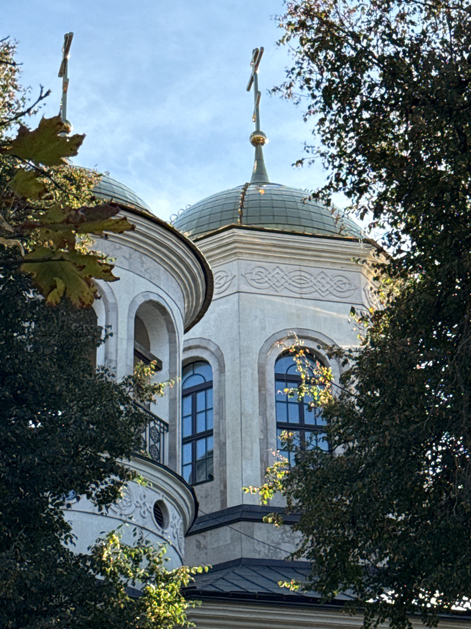 The image shows the Ascension Cathedral in Zvenigorod, Russia. The cathedral's distinctive green domes and white stone facade are visible, framed by the branches of trees. One dome features a golden cross, as does the smaller turret to the left. Arched windows with dark frames are present on the building. The sky is a clear blue with some wispy clouds. The trees have green and yellowing leaves, suggesting an autumn setting. The lighting indicates it is daytime.