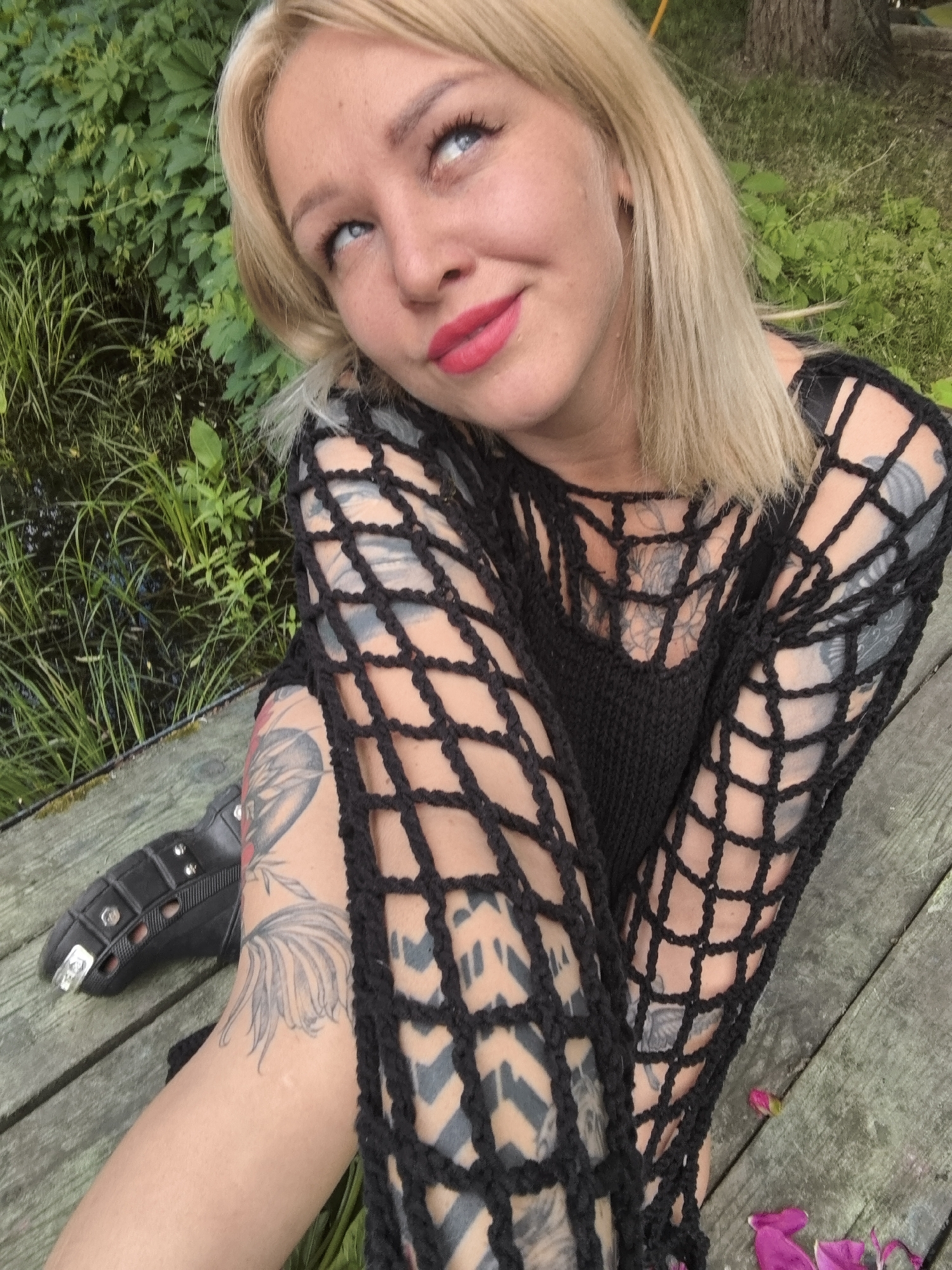 A young blonde woman with blue eyes and bright red lipstick looks up and to the left, with a slight smile. She is wearing a black, crocheted mesh top with long sleeves, revealing tattoos on her arms and chest. Her right leg is bent, and a black boot with metal studs and spikes is visible. She is sitting on a weathered wooden deck next to a body of water with tall green grass and reeds. The scene appears to be outdoors during the daytime, with lush green foliage in the background. The overall mood is casual and relaxed.