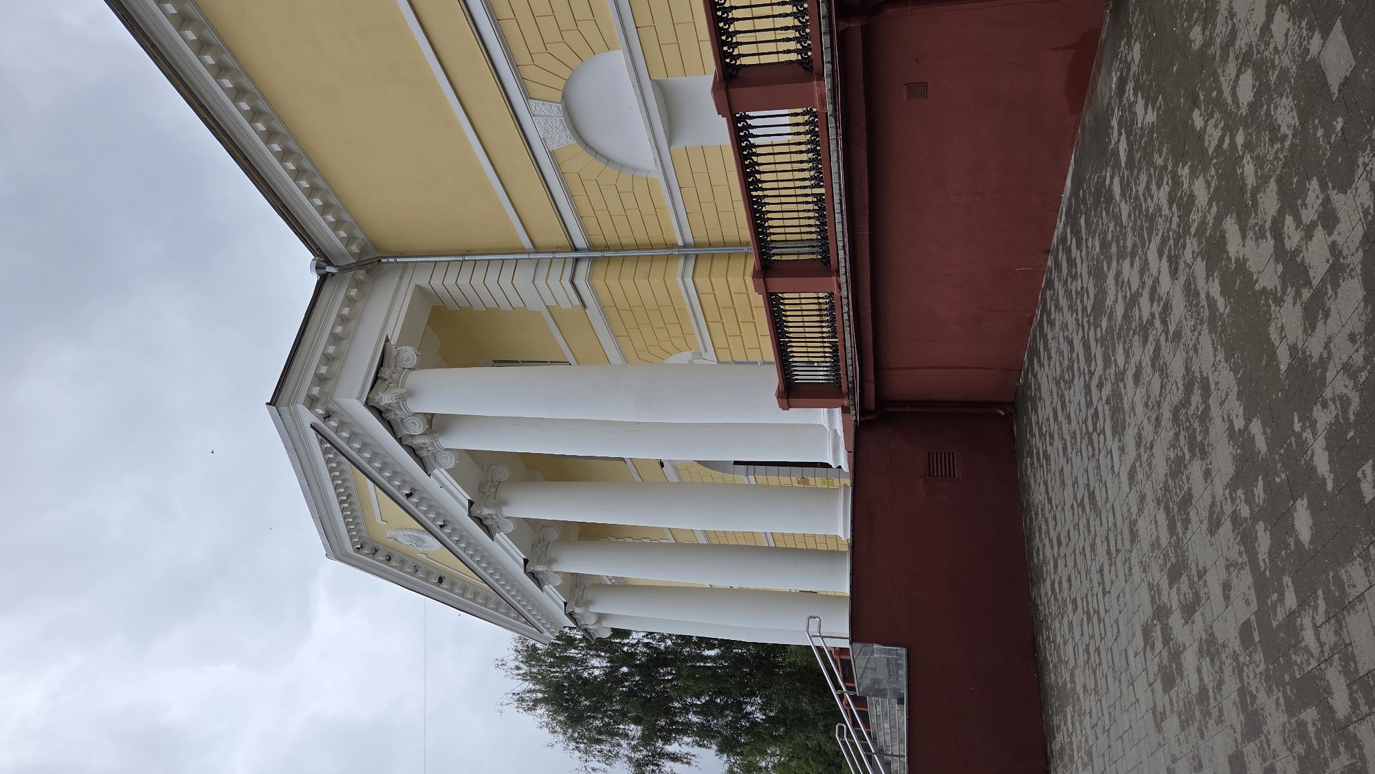 The image shows a neoclassical building with a yellow facade and a white colonnade. The building is located in the city of Vichuga, Russia. The foreground is a paved courtyard, wet from recent rain. A red retaining wall runs along the front of the building, topped with a black wrought-iron railing. A set of stairs with a metal handrail leads up to the entrance of the building, which is partially visible between the columns. A tall tree with green foliage stands to the left of the building. The sky is overcast and grey.