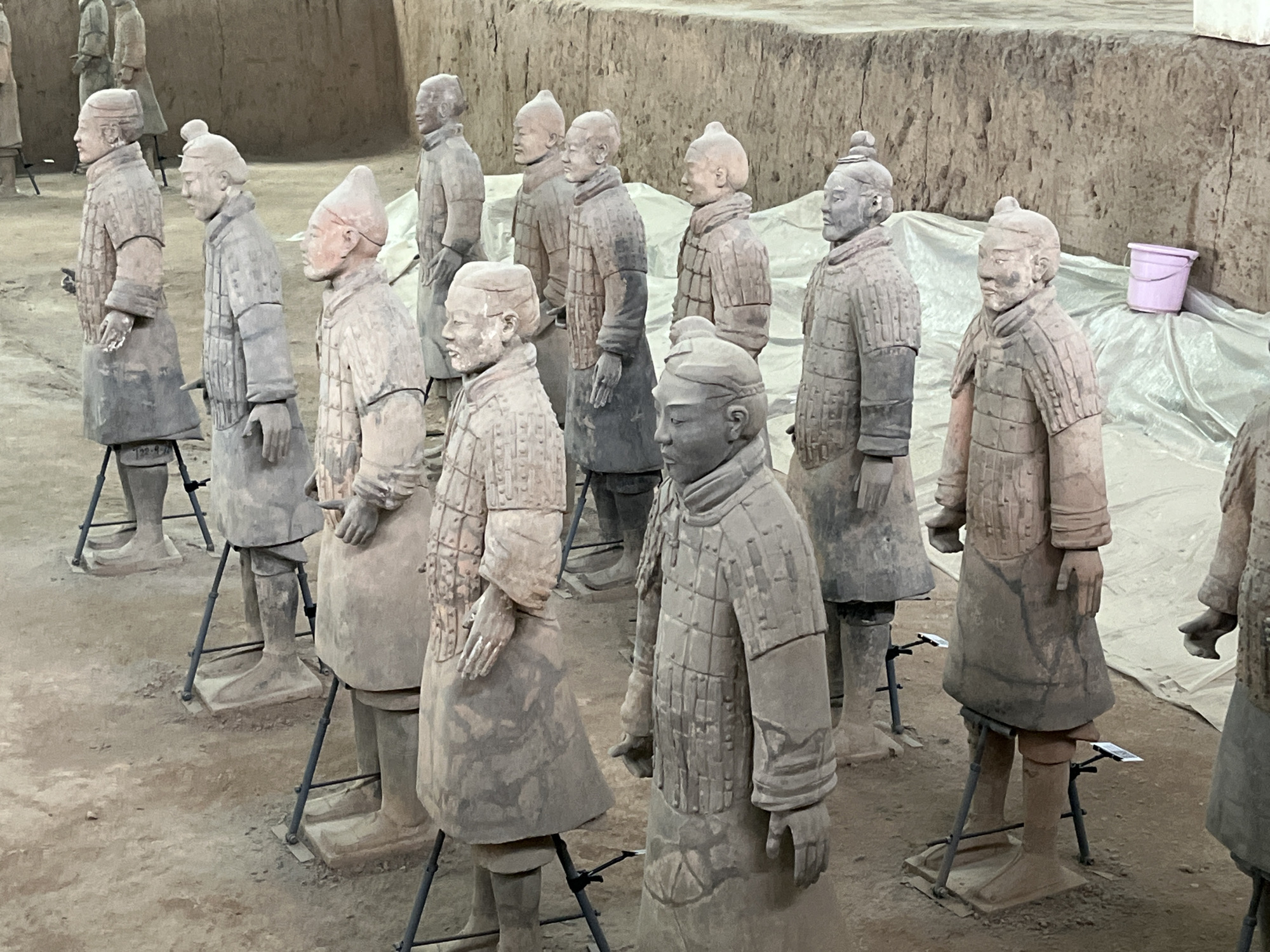 The image shows a large gathering of terracotta warrior figures, likely from the Terracotta Army in Xi'an, China. These life-sized figures are made of clay and are arranged in rows, facing forward. They are dressed in ancient Chinese military attire, including armor, helmets, and robes. The figures vary in height and pose, suggesting different ranks and roles within the army. The ground is covered in dirt and sand, and some of the figures are supported by metal stands. In the background, a large earthen wall and some protective sheeting are visible, hinting at the archaeological context of the scene. A small pink bucket is placed on the sheeting in the background. The lighting is natural, suggesting an outdoor or well-lit indoor setting.