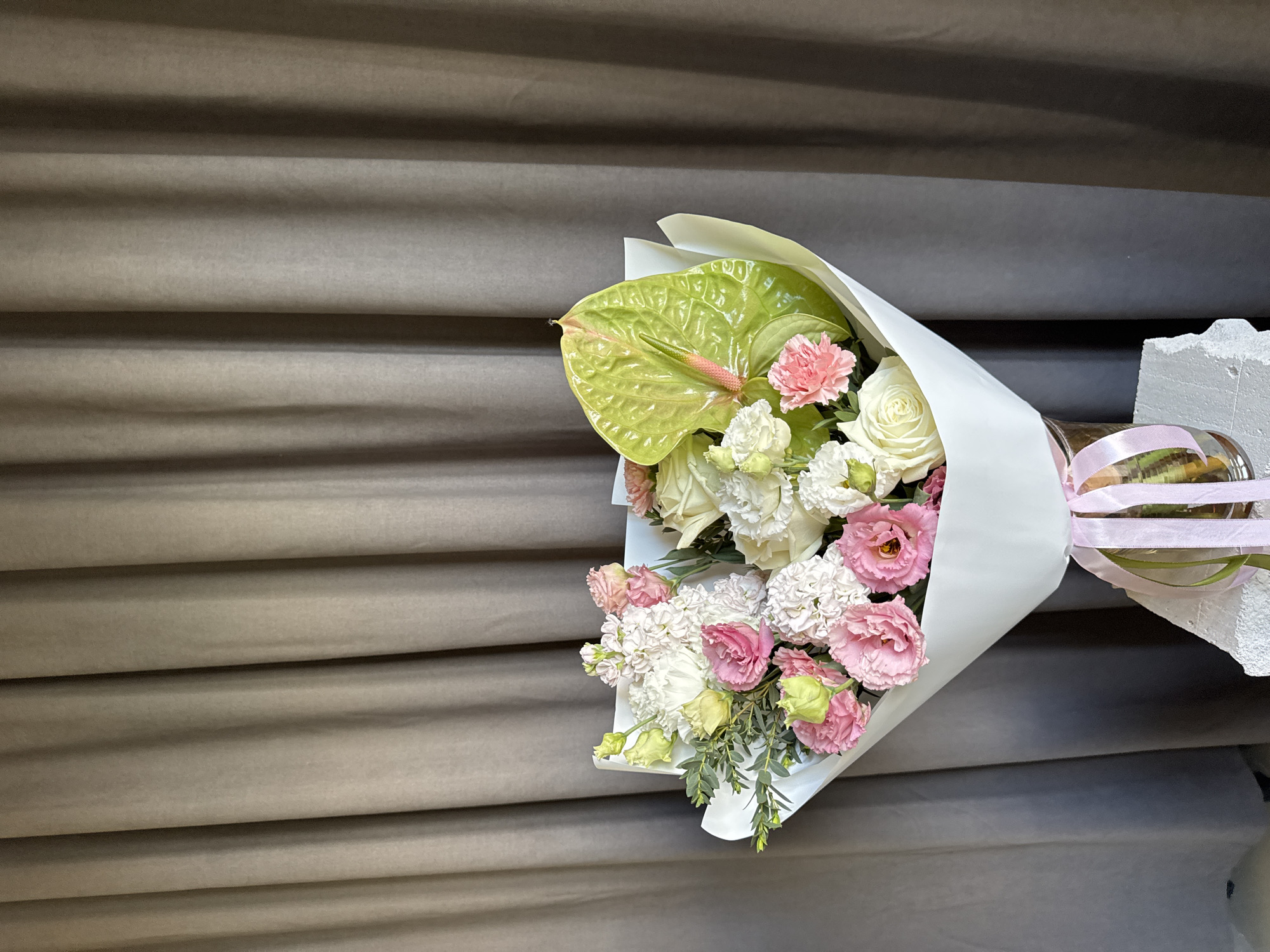 This image captures a meticulously arranged floral bouquet presented in an indoor setting within the city of Izhevsk, Russia.
The central subject is a voluminous bouquet of fresh cut flowers, predominantly featuring white and various shades of pink and light green. Specific flower types observable include white and pink Eustoma (Lisianthus), white and pink Carnations, and several white Roses. A prominent light green Anthurium, characterized by its heart-shaped spathe and pink spadix, is positioned on the right side of the bouquet. Greenery, likely Eucalyptus, is interspersed throughout the arrangement. The bouquet is wrapped in pristine white floral paper, gathered at the bottom to form a conical base. Multiple strands of light pink satin ribbon are tied around the wrapped base, cascading downwards.
The wrapped bouquet is situated in a clear glass vase, which rests upon a rough, off-white, block-like stand, possibly made of concrete or plaster, featuring an irregular, textured surface on its visible edge.
The background consists of a uniformly draped wall of heavy, taupe-colored curtains. These curtains display prominent vertical pleats and folds, indicating a substantial fabric. The lighting is soft and diffused, illuminating the scene evenly without harsh shadows. No individuals are present in the image.