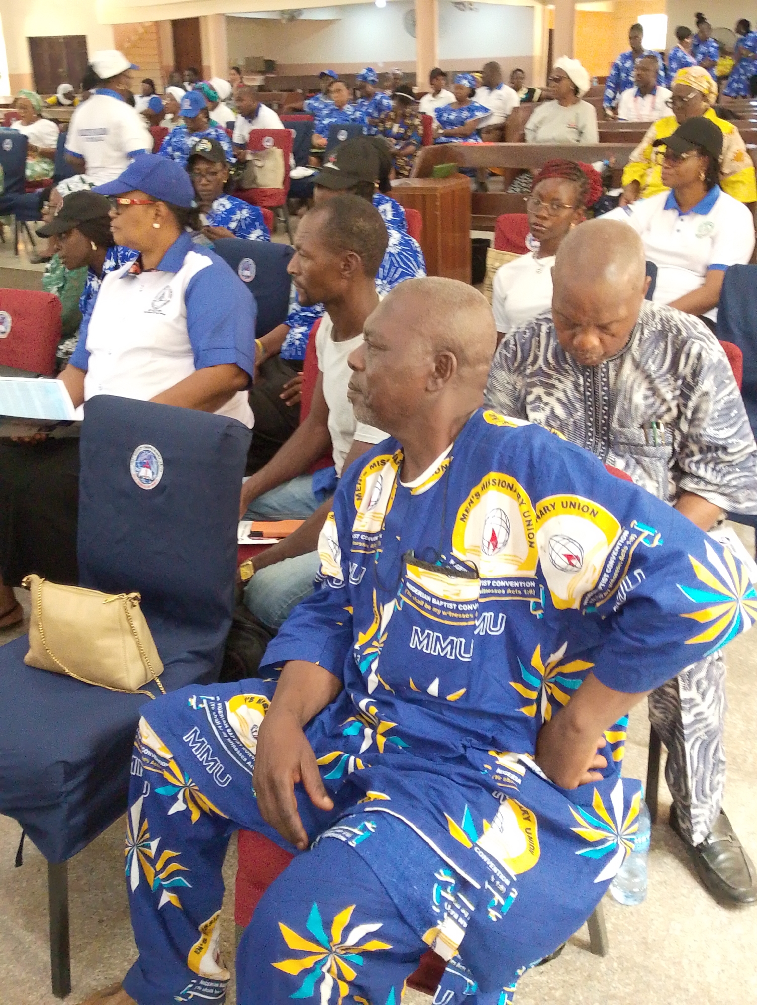 This image depicts an indoor gathering of numerous adults, likely a convention or meeting, within a hall or large room in Kubwa, Nigeria, during the daytime. The attendees are predominantly dressed in matching blue and white attire, indicating affiliation with a specific organization. Many wear white polo shirts with blue sleeves and blue caps, while others sport blue patterned outfits.
Visible text on the clothing, particularly on a man in the foreground wearing a distinct blue and yellow patterned suit, identifies the event as related to the "Men's Missionary Union" (MMU) and the "Nigerian Baptist Convention." His outfit also features the scripture "Ye shall be my witnesses," likely from Acts 1:8.
Participants are seated in rows of chairs; some are blue with a circular logo, others are red. In the background, wooden benches are also visible. A woman on the left in the blue and white uniform holds a document, and a light brown handbag rests on an empty blue chair beside her. People appear attentive and calm, observing the proceedings of the event. Water bottles are scattered on the floor, suggesting a lengthy gathering. The setting implies a formal, organized assembly, possibly a religious conference or convention.