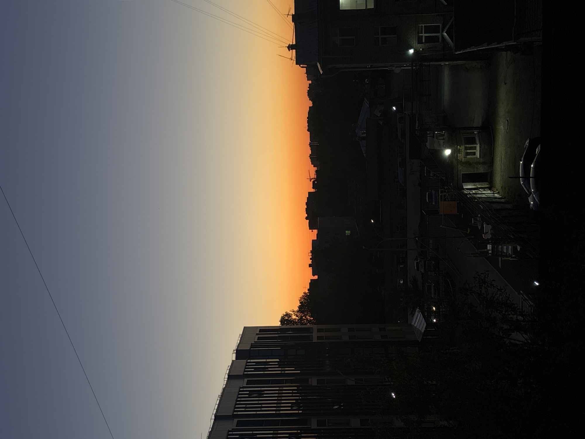 The image shows a cityscape at dusk in Perm, Russia. The sky transitions from a pale blue to a vibrant orange, indicating the setting sun. Silhouettes of buildings and trees form a dark outline against the colorful sky. One building is modern with horizontal lines. Other buildings and structures are dense and have lit windows, suggesting residential or commercial areas. Streetlights illuminate parts of a street and parking lot, where cars are visible. A few power lines cross the sky.
