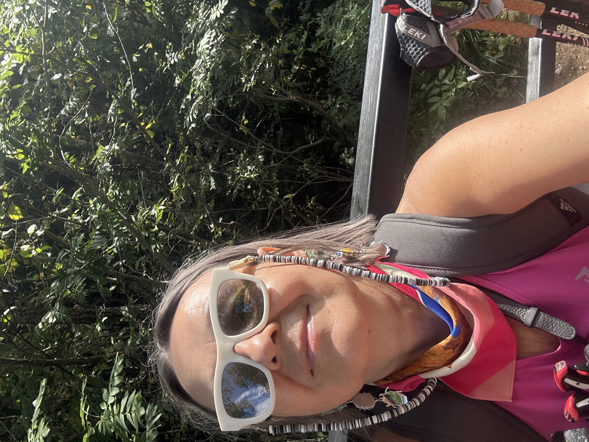 A woman with gray hair and white-rimmed sunglasses takes a selfie outdoors, with lush green foliage in the background. She is wearing a pink tank top and a colorful patterned scarf around her neck. A beaded chain with charms hangs from her sunglasses. To the right, a pair of trekking poles with "LEKI" branding are visible. The scene appears to be a park or natural area.