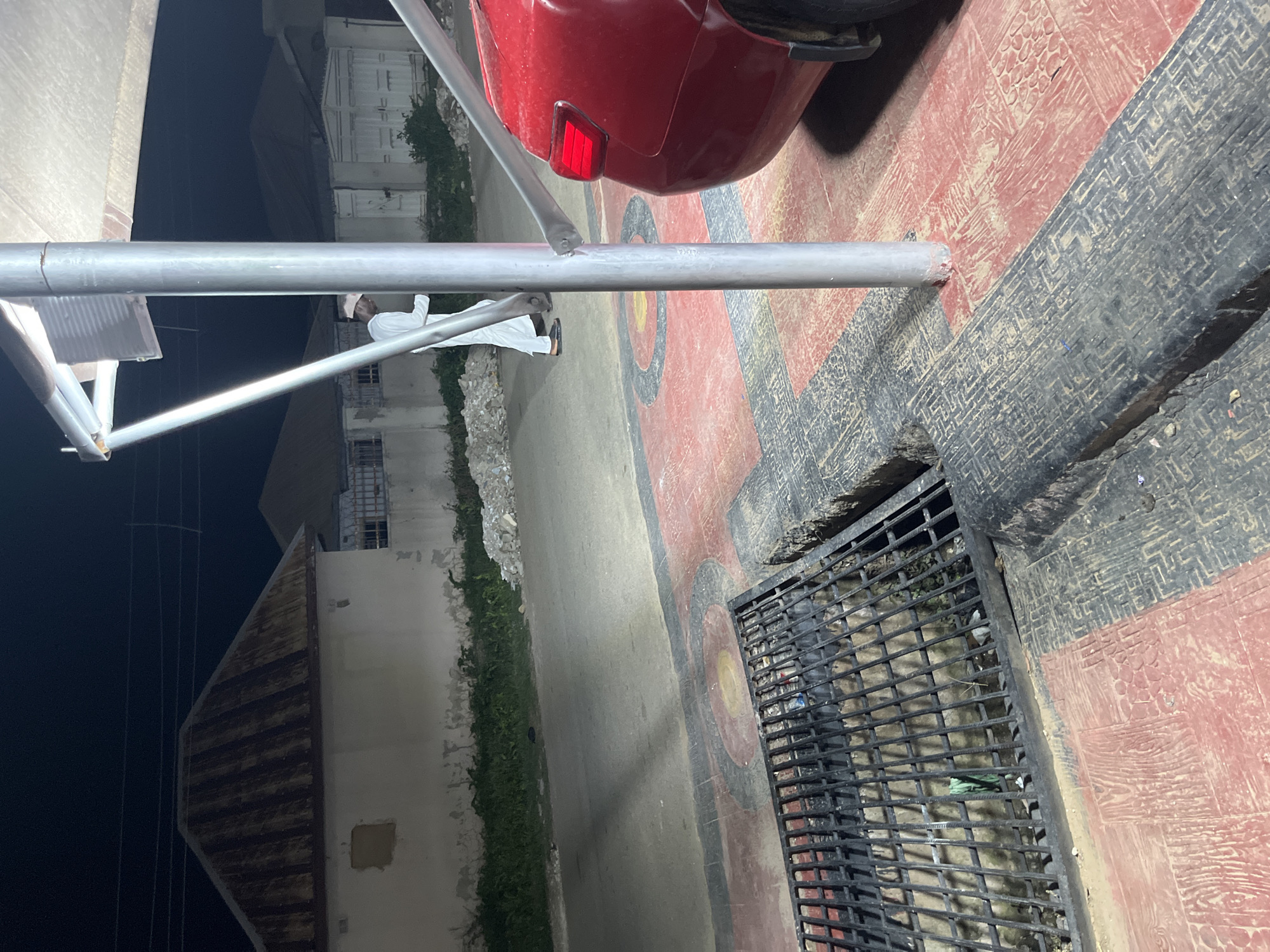 In the city of Abuja, Nigeria, at night, a man in a white robe and cap walks past a red car. He is partially obscured by a metal pole supporting a canopy. The man is walking away from the camera, his face not visible. The environment is a paved area with red and black patterned tiles, a metal grate over a drain, and some scattered debris. In the background, houses with corrugated metal roofs are visible, along with a street and some overgrown vegetation. The lighting suggests artificial illumination from the canopy and possibly streetlights.