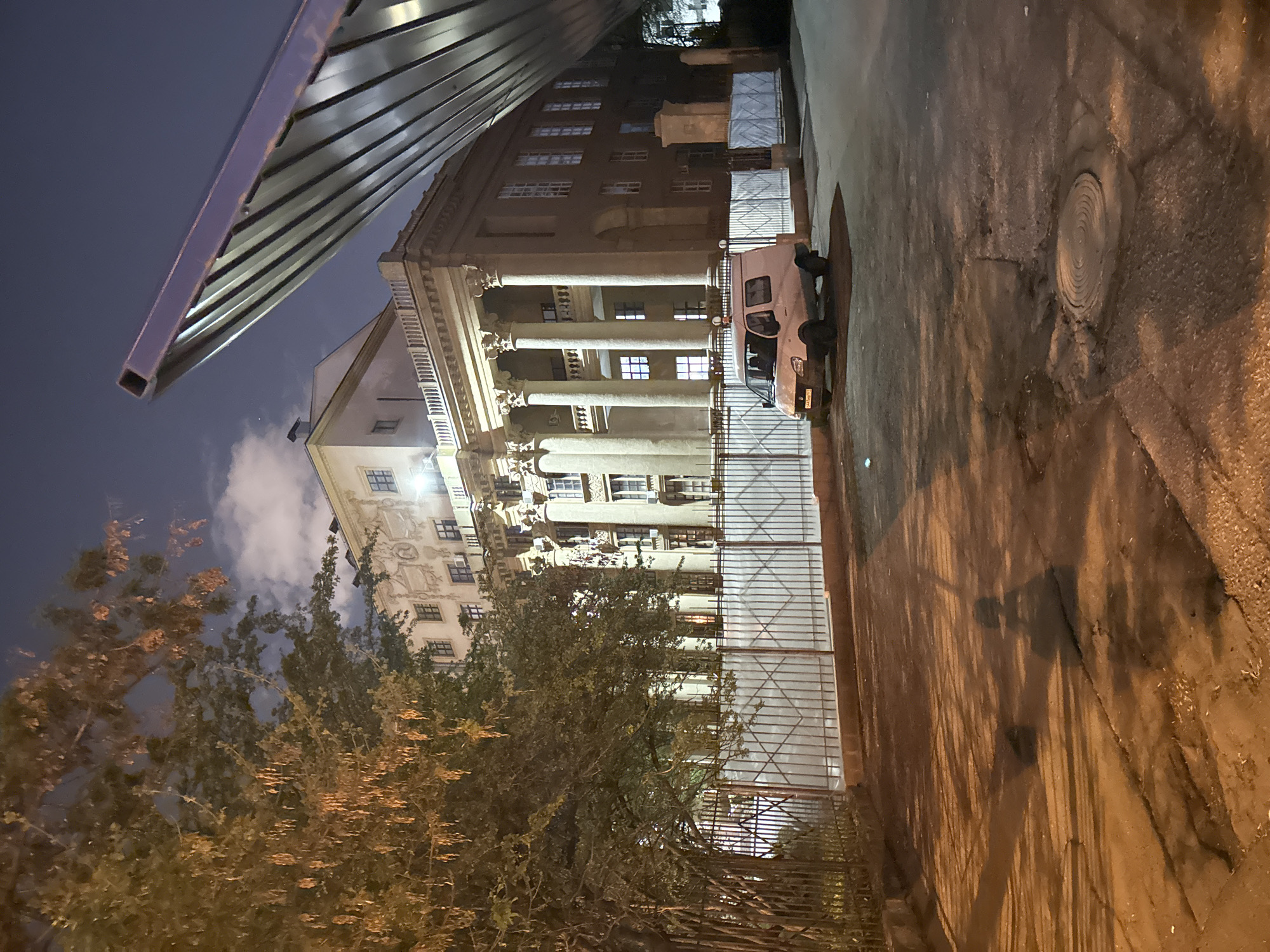The image depicts a street scene in Moscow, Russia, at night. A large building with classical architectural elements, including columns, is visible behind a metal fence. A white van is parked alongside the fence. A tree with autumnal foliage is in the foreground. A metal canopy or structure is partially visible in the upper left of the image. The street is paved and has a manhole cover. The lighting is dim, suggesting it is either late evening or early morning.