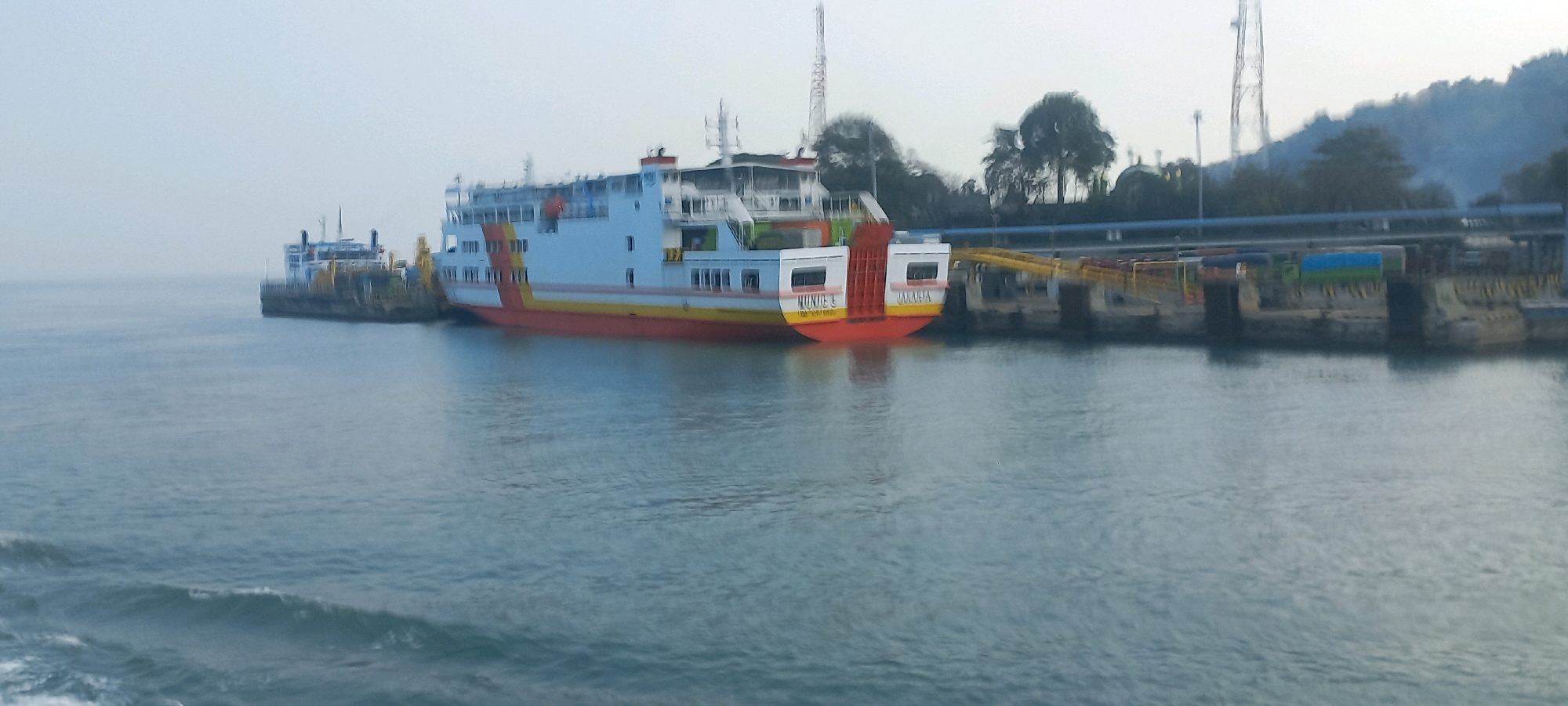 A wide-angle, slightly hazy, daylight shot captures a ferry terminal in Cilegon, Indonesia, with two large ferries docked. The primary ferry, a passenger and vehicle ferry, is prominently displayed, painted white with orange and yellow stripes along its hull. It is docked at a pier constructed with yellow metal ramps, connecting to a concrete embankment. The ferry has multiple decks with visible windows and railings. The name "MUNICE" is visible on the stern, along with the word "JAKARTA." A second, smaller ferry is visible docked to the left of the main ferry, appearing to be a similar vessel.
The water in the foreground is a calm, grey-blue, with some minor ripples and a faint wake on the left side, suggesting the photographer is on another moving vessel. The background shows a densely forested hillside under a pale, overcast sky. A tall, lattice-like communication tower is visible on the hillside behind the ferry terminal. There are trees and some structures along the shoreline. The overall impression is one of a busy port environment on an overcast day. No people are distinctly visible on the ferries or the dock.