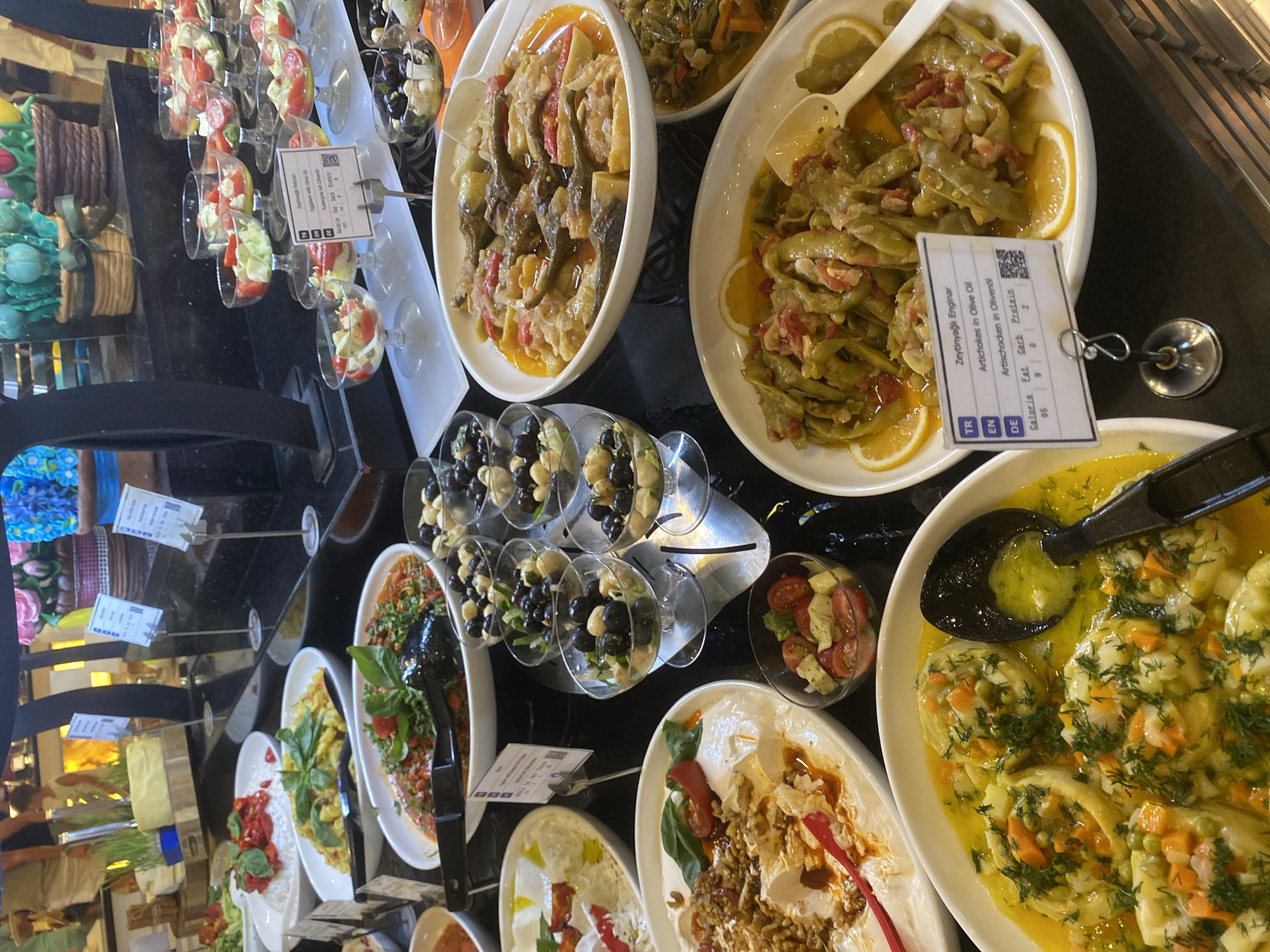 The image shows a buffet-style food display at a hotel in Aksu, Turkey. The display features a variety of dishes, including salads, vegetable stews, and dips. Many of the dishes are served in white ceramic bowls, with some in glass cups. There are also several small signs in front of the dishes, providing information about the ingredients and nutritional content. In the background, blurred figures of people can be seen browsing the buffet. The lighting is bright, suggesting an indoor setting.