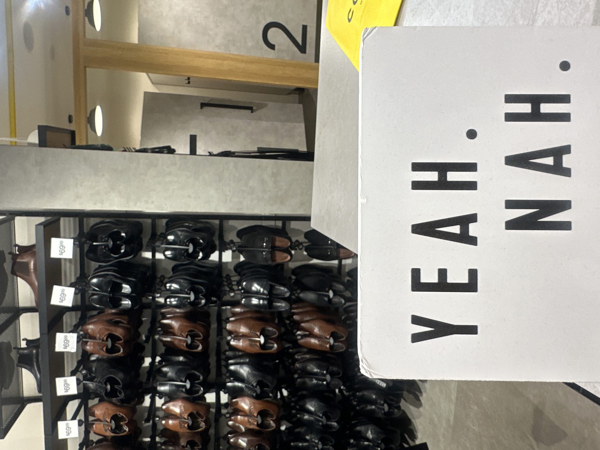 This image shows a retail store interior, likely a shoe store, with shelves displaying numerous pairs of men's dress shoes and boots. The shoes are arranged in rows and appear to be of various styles and colors, predominantly black and brown. Some shoes have price tags with "$69.99" visible. In the foreground, a white sign with black lettering reads "YEAH. NAH." This phrase is a colloquial Australian expression of polite refusal or disagreement. In the background, there are changing rooms with doors, and one changing room is marked with the number "2". The overall environment suggests a commercial setting with good lighting. There are no people visible in the image. The mention of "Coffs Harbour, Australia" suggests this scene is likely a retail establishment within that city.
