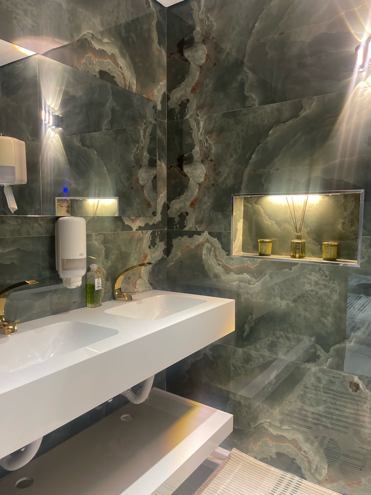 This image depicts a modern bathroom with a marble-like green tiled wall. A white countertop with two sinks is installed, featuring gold-colored faucets. To the left of the sinks, a white soap dispenser is mounted on the wall. Next to the soap dispenser, on the countertop, is a clear glass bottle with green liquid, possibly hand sanitizer or soap. In the background, a recessed shelf is illuminated by warm light, holding a diffuser with reeds and two small jars. The lighting in the bathroom is a mix of overhead and recessed fixtures, creating reflections on the shiny surfaces. The overall aesthetic is sleek and luxurious. The location is identified as Jabriya, Kuwait.