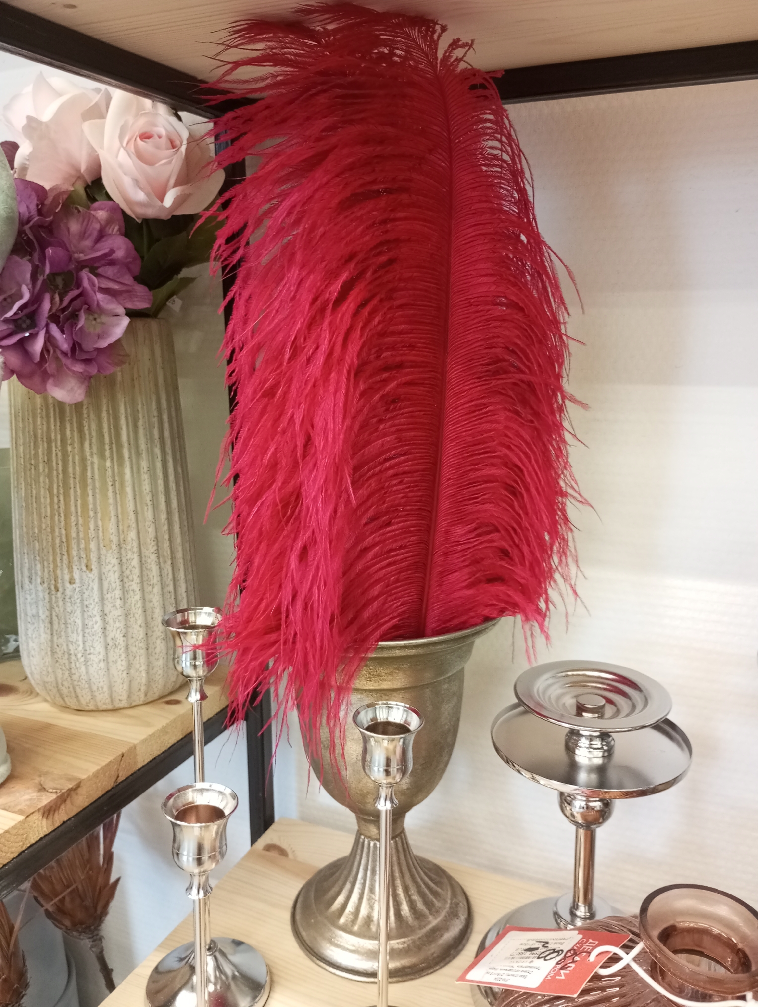 The image shows a shelf display, likely in a retail setting in Izhevsk, Russia. The focus is on decorative items, including: a vase containing pink and purple artificial flowers, a metallic urn holding large red feathers, two silver candlesticks, and a silver tiered stand. There is a price tag in Russian visible near the base of the tiered stand. The scene is well-lit, suggesting an indoor environment.
