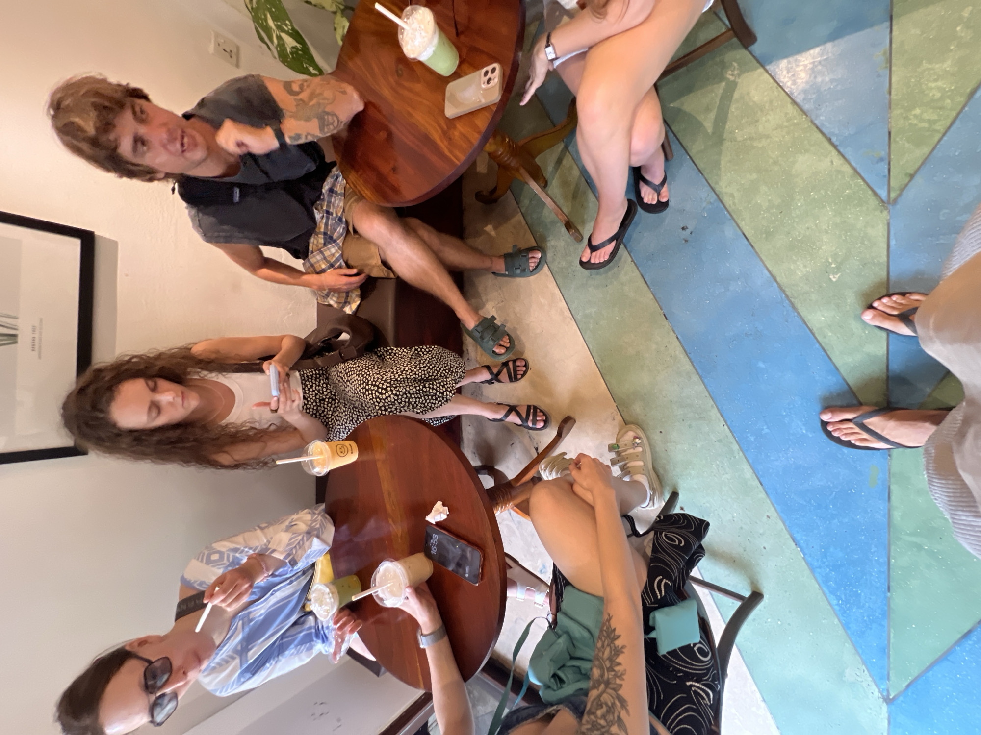 The image depicts a group of four individuals seated around a circular wooden table in what appears to be a cafe or restaurant. The floor has a distinctive pattern of intersecting blue and green geometric shapes. In the foreground, two feet wearing flip-flops are visible, suggesting the photo is taken from the perspective of someone standing.
On the table, there are several drinks: two green beverages in clear cups with straws, a lighter colored drink in a clear cup with a straw, and a yellow drink in a clear cup with a straw and a smiley face sticker. A mobile phone displaying the time 16:53 is also on the table.
The individuals are dressed casually. One person, on the left, wears a blue and white striped shirt and sunglasses, and is eating. Another person, in the center, has curly hair, wears a white tank top and a black and white patterned skirt, and is looking at their phone. To the right, a person with brown hair wears a dark t-shirt and plaid shorts, and is looking towards the camera. Another person, partially visible on the far right, is wearing a white top and has their legs crossed, also wearing sandals. One individual has a tattoo on their arm.
The environment suggests an indoor setting with a white wall and a framed picture on the wall in the background. A plant with large leaves is visible on the right side of the image.
While the image does not contain explicit visual cues indicating the city of Galle, Sri Lanka, the vibrant, geometric floor pattern is reminiscent of contemporary design found in some cafes and public spaces in tourist areas of cities like Galle, which often incorporate modern aesthetics with a local flair.