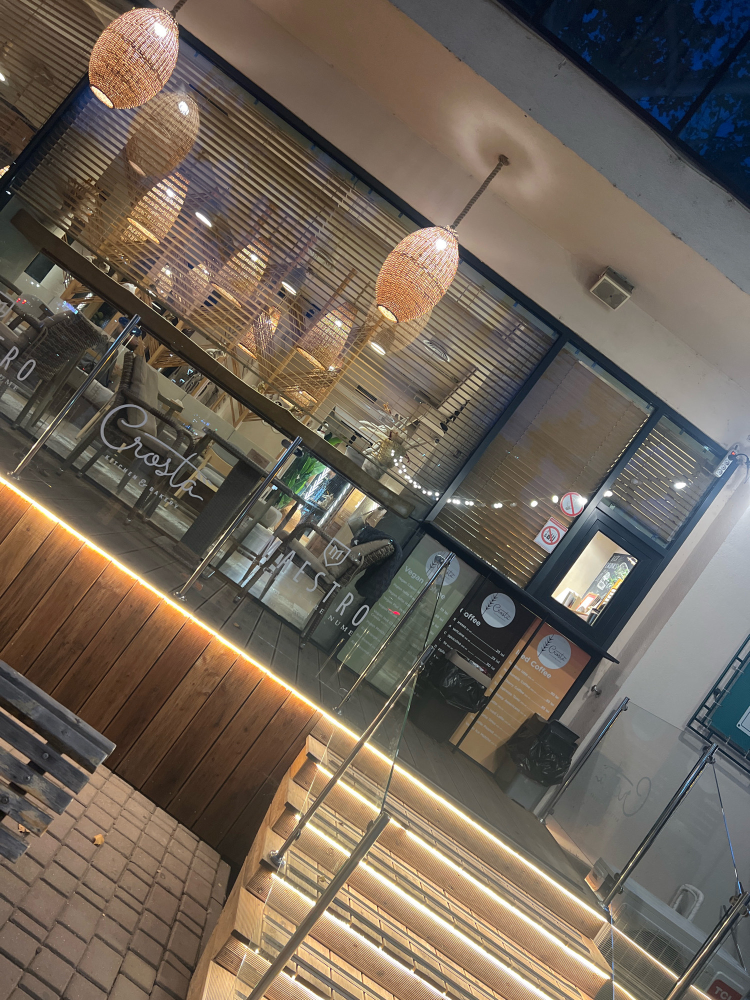 The image shows the exterior of a cafe at night. The cafe has a modern design with large glass windows and wooden accents. Inside, tables and chairs are visible, along with decorative lighting. The cafe's name, "Crosta Kitchen & Bakery," is displayed on the glass. The exterior features a wooden deck area with a bench and paved ground. The lighting suggests it is evening or nighttime. There are no people visible in the image.
