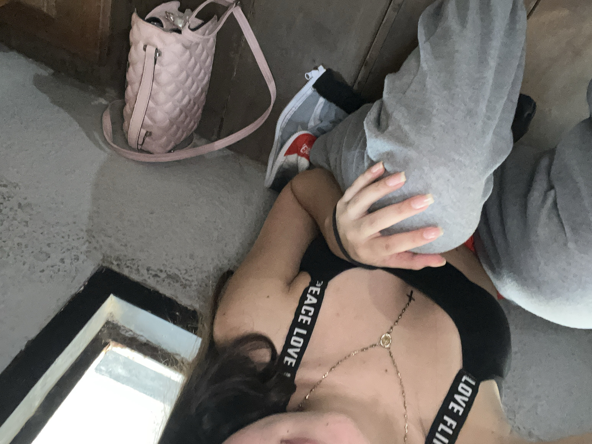 The image presents an overhead view of a person, likely a woman, reclining indoors in a casual setting. The woman is visible from her upper torso downwards, wearing grey sweatpants and a black top or sports bra. A gold body chain, featuring a small circular pendant and a cross, drapes across her chest. Her left hand, adorned with long, light-colored fingernails, rests on her leg. Dark hair frames the left side of her face, which is mostly cropped out of the frame.
To the upper left, a light pink quilted bucket bag with a long strap is placed on a muted grey surface, appearing slightly open. Near the woman's legs, a red sneaker, possibly Adidas given a visible logo, peeks out from an open white and black zippered bag or container.
Notable text, "PEACE LOVE", is clearly visible on the strap of her black top. The remainder of the text on the strap is partially obscured but appears to continue with another word. In the bottom left corner, a dark-framed, light-colored architectural detail, resembling a window or an alcove, is visible, suggesting a source of natural light.
The scene conveys a relaxed and informal atmosphere, likely a personal moment or selfie. The overall lighting indicates it is daytime. The provided location context is Juan Griego, Venezuela.