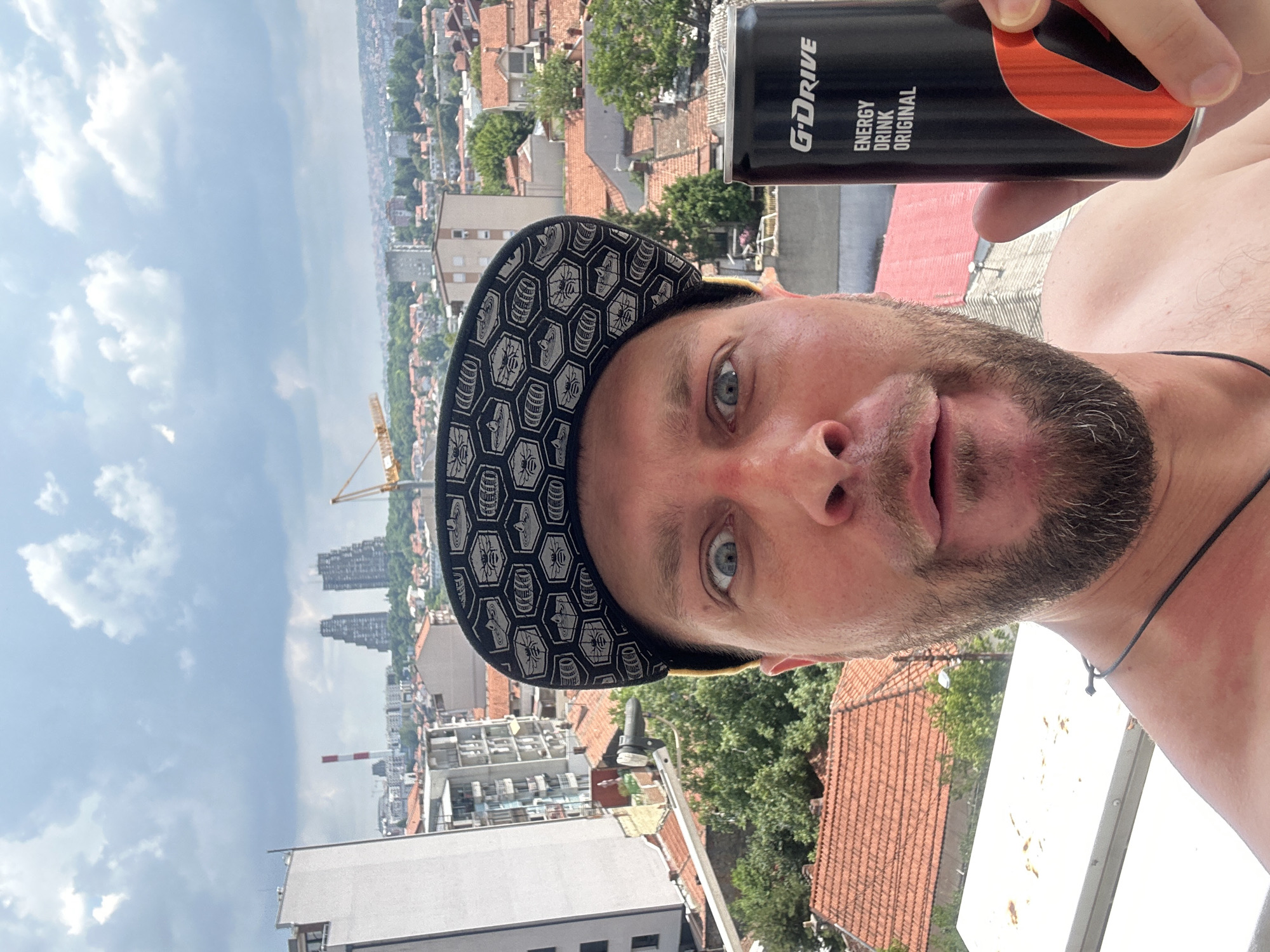 A man in a black baseball cap with a hexagonal pattern and a yellow brim is taking a selfie. He is holding a black can of "G-DRIVE ENERGY DRINK ORIGINAL" in his right hand. The man has a beard and light-colored eyes. He is shirtless. The background shows a cityscape with buildings, red-tiled roofs, green trees, and a cloudy sky. In the distance, two tall, angular buildings and a construction crane are visible, along with a red and white striped smokestack. This cityscape is consistent with the Zvezdara Urban Municipality in Serbia, known for its mix of residential buildings, greenery, and industrial elements. The image appears to have been taken from a balcony or elevated viewpoint.
