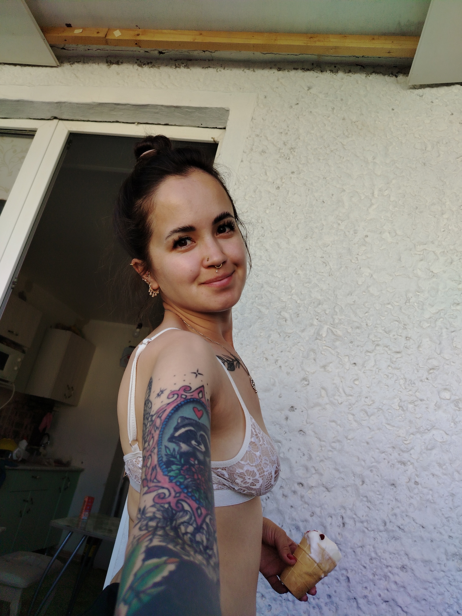 The media file shows a young woman in a white lace bralette, holding an ice cream cone. She has dark hair tied up in a bun and is wearing hoop earrings and a nose ring. Her left arm is covered in colorful tattoos, including a raccoon in a frame. She is smiling at the camera, and the overall mood appears relaxed and happy.
The setting appears to be outdoors, possibly on a balcony or patio, with a textured white wall in the background. To the left, a doorway leads into what looks like a kitchen or dining area, with cabinets and a microwave visible. The time of day appears to be daytime, judging by the lighting. The location is identified as Noviy, Russia. There is no visible text in the image.