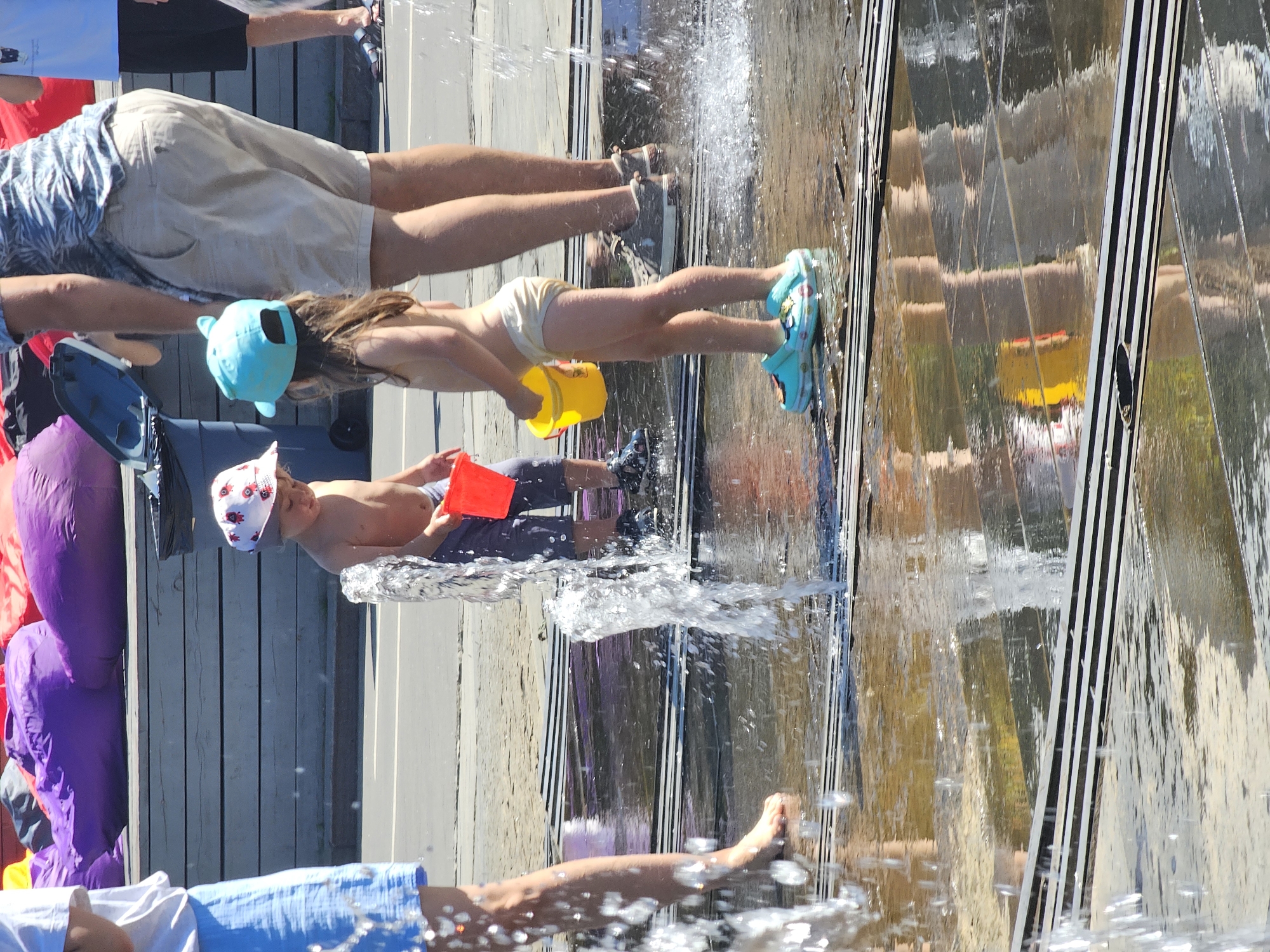The image shows a group of people, including children, playing in a water feature. The sun is shining brightly, indicating it is daytime, and the scene appears to be outdoors, possibly a park or public space. The water feature itself is made of reflective surfaces, creating a shimmering effect as the water cascades down.
One child, wearing a blue hat and a yellow bucket, is splashing in the water. Another child, wearing a white cap with red ladybug-like designs, is holding a red bucket and is partially submerged in the water. Several adults are also present, observing or participating in the activity. Their legs are visible, wearing shorts and various footwear. The background shows wooden planks, suggesting a boardwalk or a similar structure.
The atmosphere seems to be cheerful and playful, with the water and bright sunlight contributing to a summery, recreational feel. There is no visible text in the image. The location context of Moscow, Russia, suggests this is likely a summer activity in a city park or public square with interactive water installations.