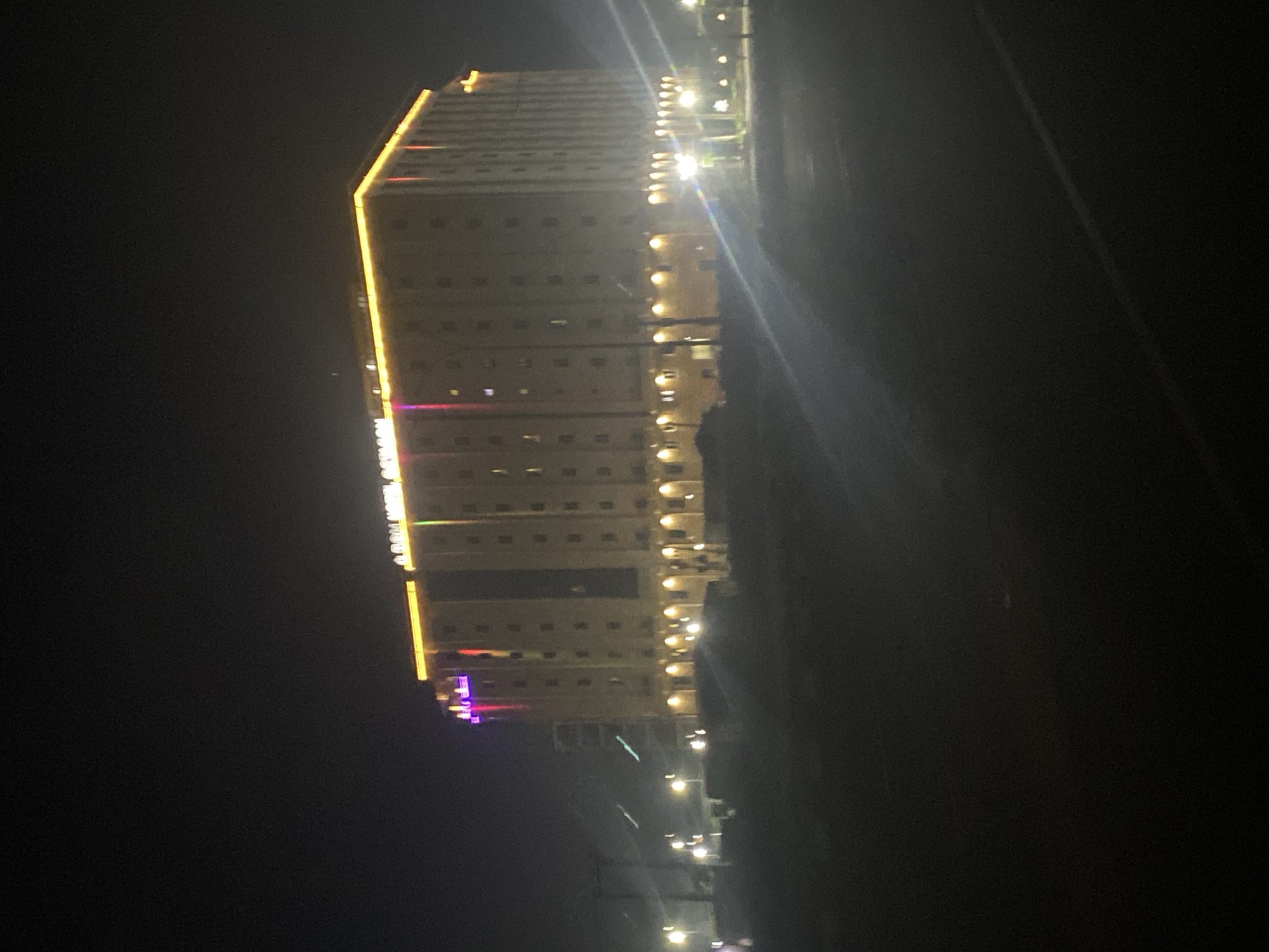 A night time, eye-level, medium shot shows a large, modern, multi-story building illuminated with colorful lights. The building appears to be a hotel, with a prominent sign that reads "ARAH HOTEL GOMBE" in bright yellow neon lights. The building itself is a light brown color and has numerous windows. The ground level of the building features arched entrances and windows, also illuminated by warm yellow lights.
In front of the building, there is a dark, open area that could be a parking lot or a plaza. Several streetlights are visible in the foreground and mid-ground, casting pools of light on the dark ground. Some of these lights are blurred, suggesting movement or a shallow depth of field. In the far distance, more lights are visible, indicating the presence of other buildings or urban infrastructure.
The sky is dark, with no stars or moon visible. There is a hazy or foggy atmosphere, which softens the edges of the buildings and lights, giving the scene a slightly ethereal quality. The overall impression is that of a well-lit, contemporary establishment in a somewhat remote or less densely populated part of the city at night. The location is specified as Jiru, Nigeria.